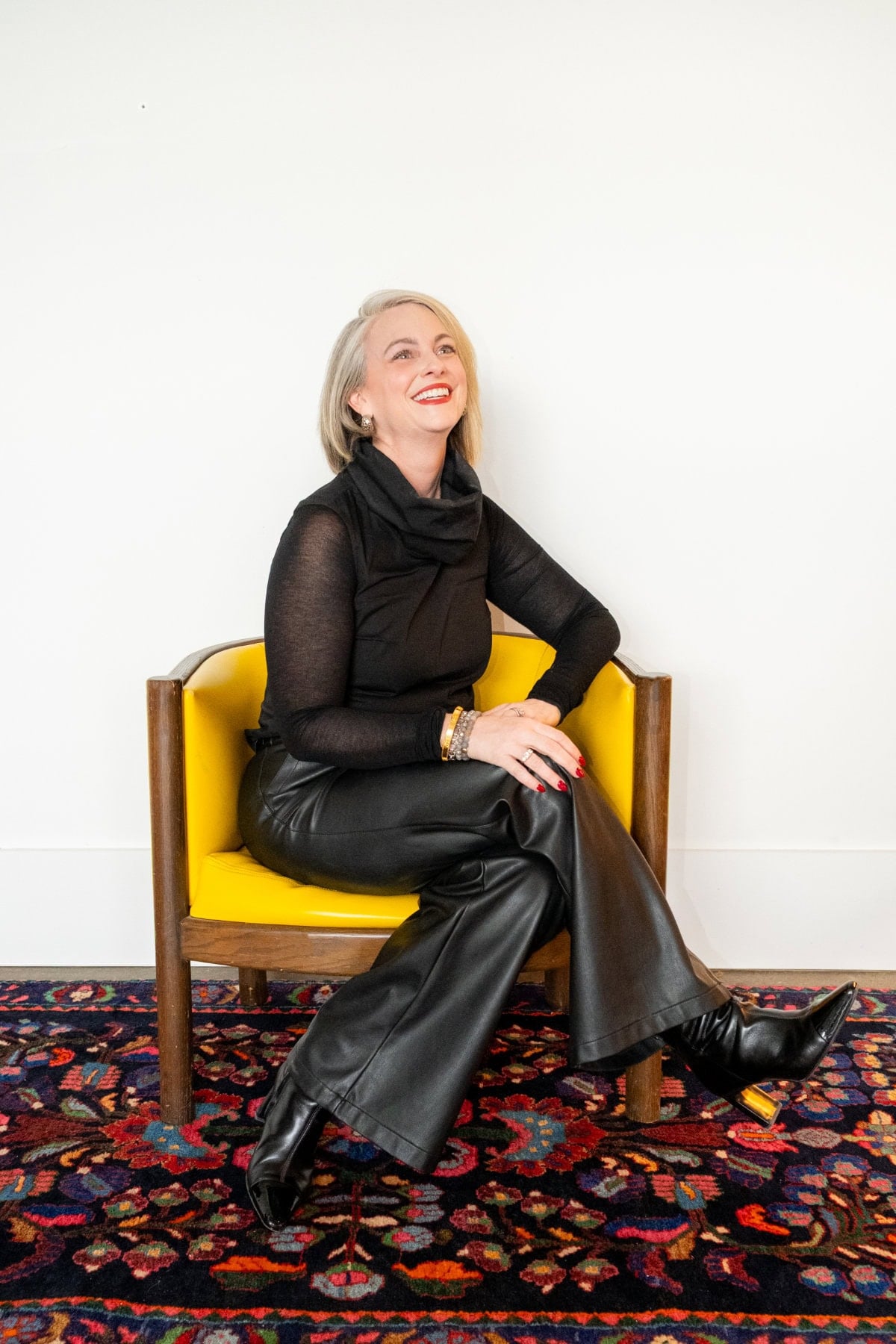 A woman with short blonde hair, dressed in black, sits smiling on a yellow chair against a white wall and patterned rug.