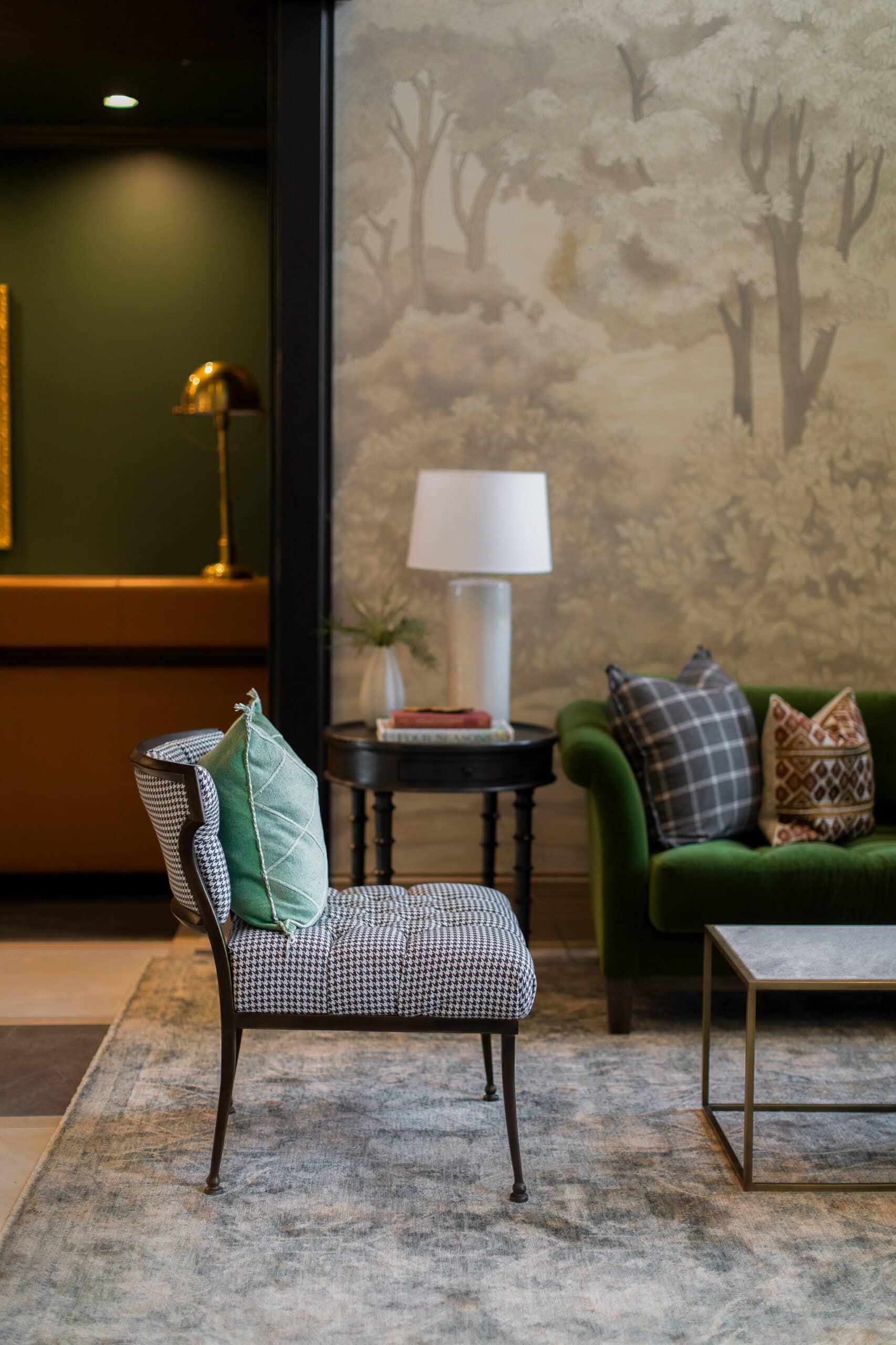 A modern living room features a patterned chair with green and gray pillows, a green sofa, a side table with a lamp, and a coffee table on a rug with a tree mural in the background.