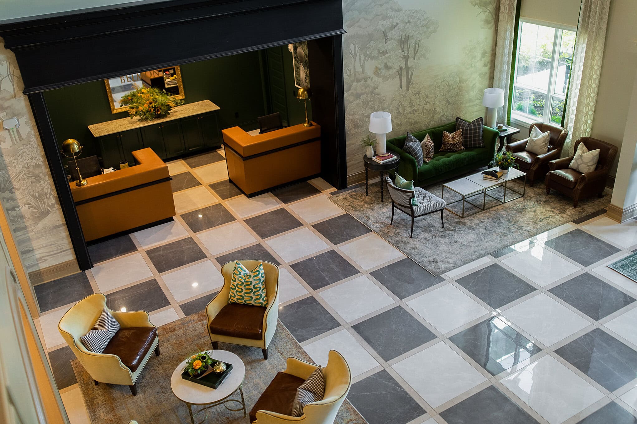 A spacious lobby with a checkered tile floor, reception desks, seating areas with chairs and a green sofa, large windows, and neutral-colored decor.