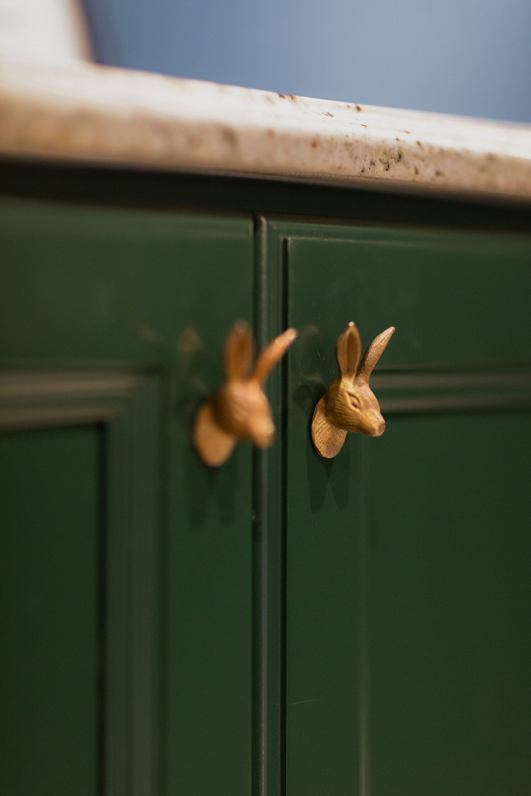 Two green cabinet doors with gold rabbit head-shaped handles and a light-colored marble countertop above.