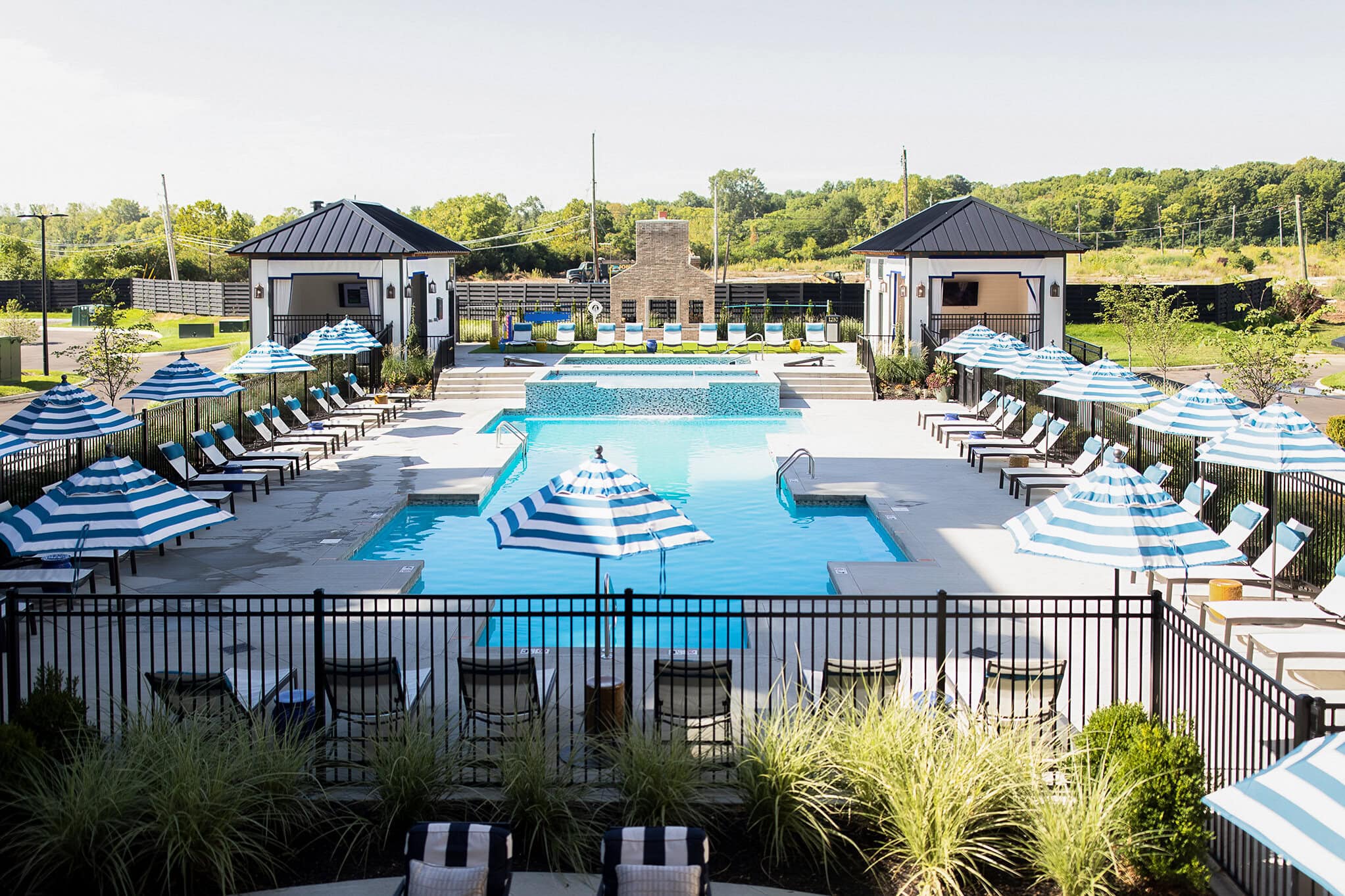 Fenced outdoor pool area with blue and white striped umbrellas, lounge chairs, two cabanas, and a central swimming pool surrounded by greenery.