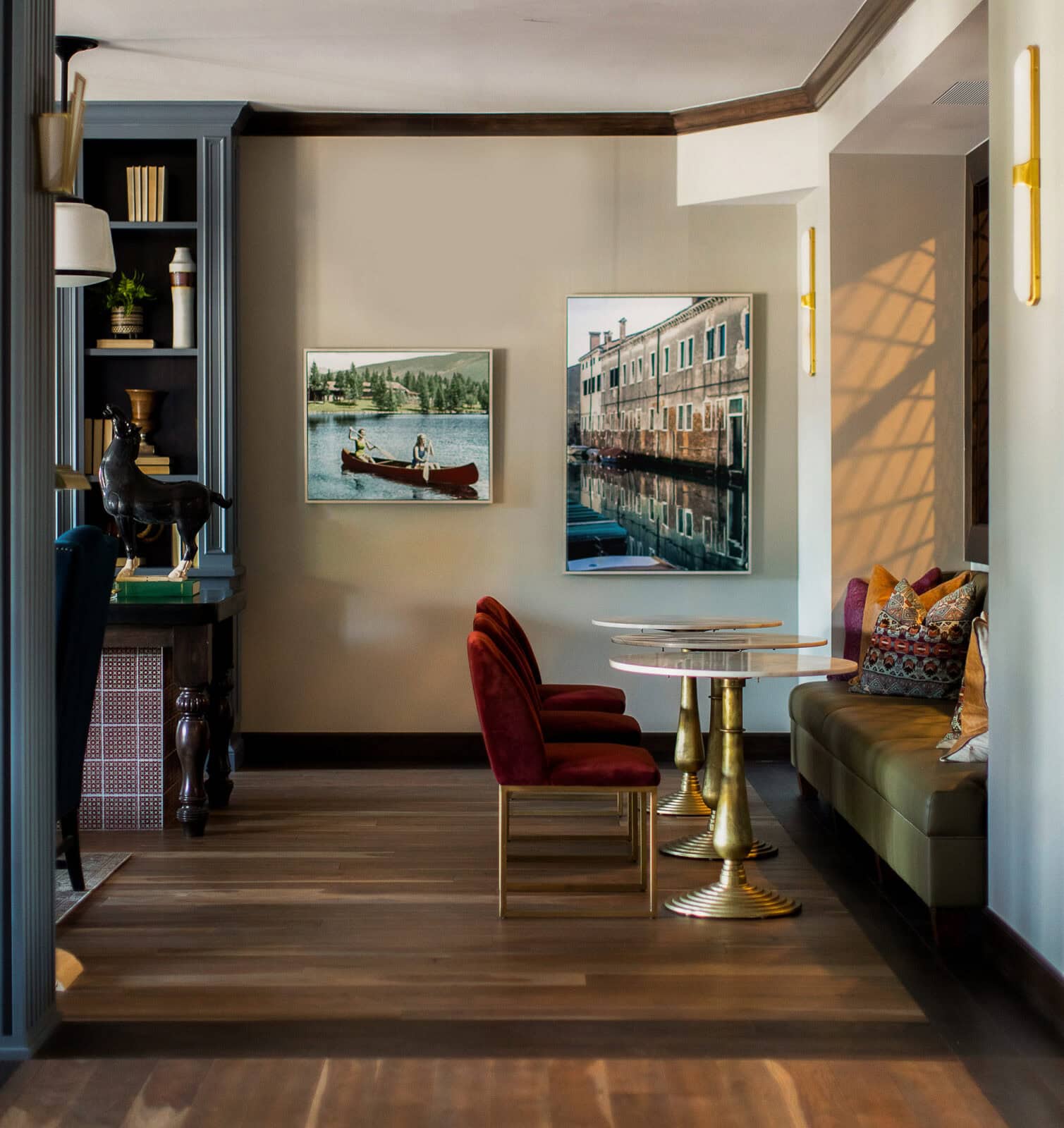 A cozy seating area with velvet chairs, a green banquette, marble tables, wall art, and wooden floors in a softly lit room.