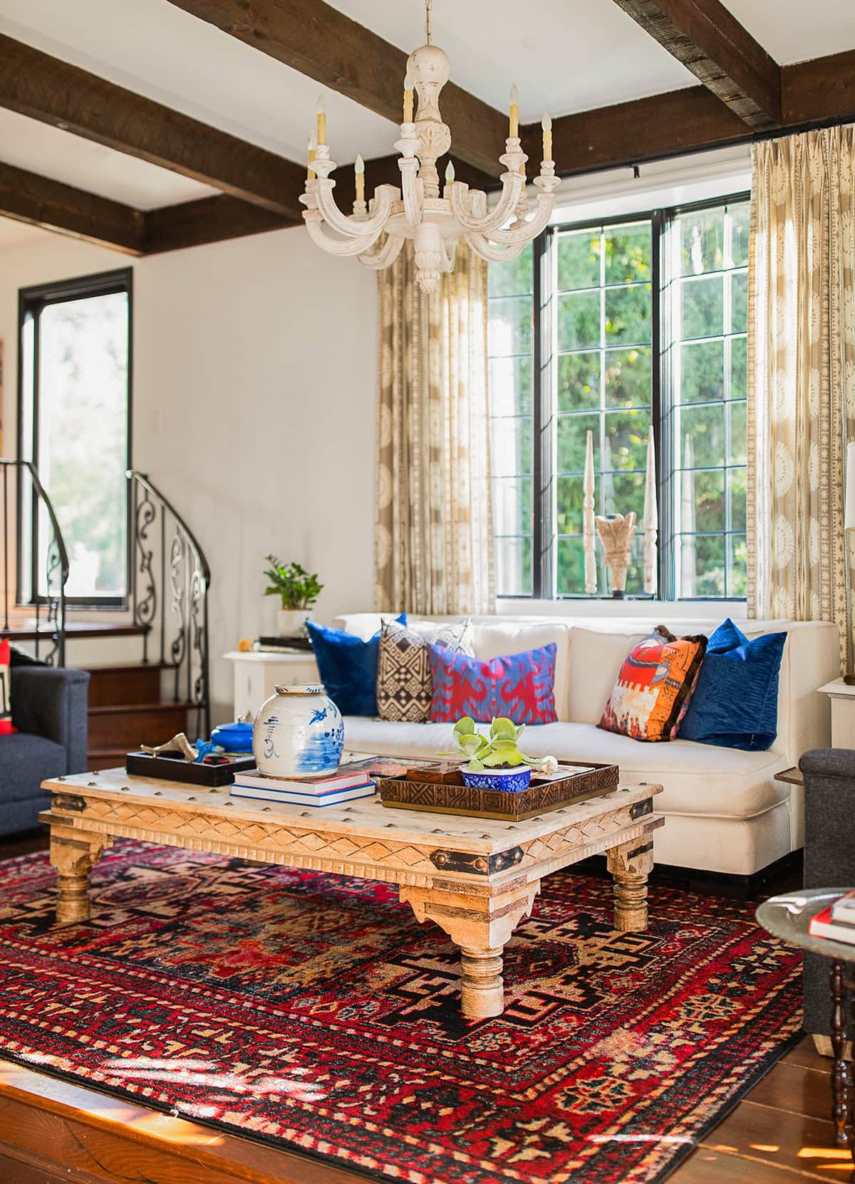 A living room with a white sofa, colorful cushions, a wooden coffee table, a red patterned rug, a white chandelier, and large windows with beige curtains.