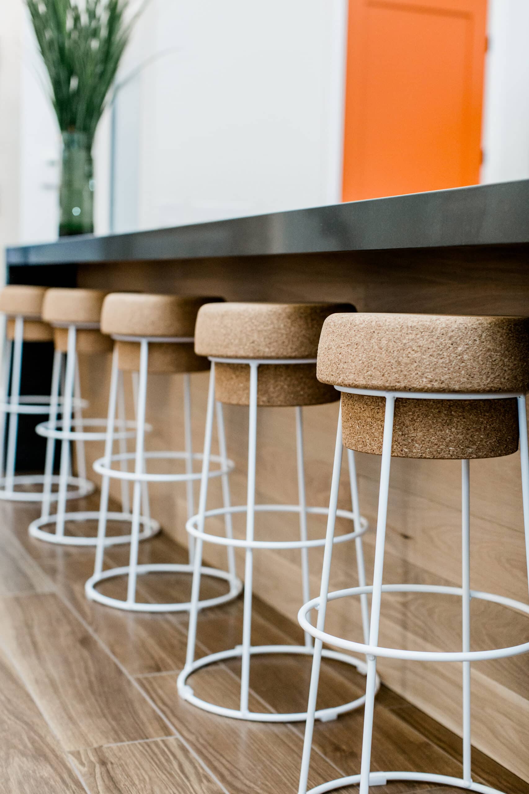 Four modern bar stools with cork seats and white metal legs lined up at a wooden counter; an orange door and a vase of tall green plants are visible in the background.