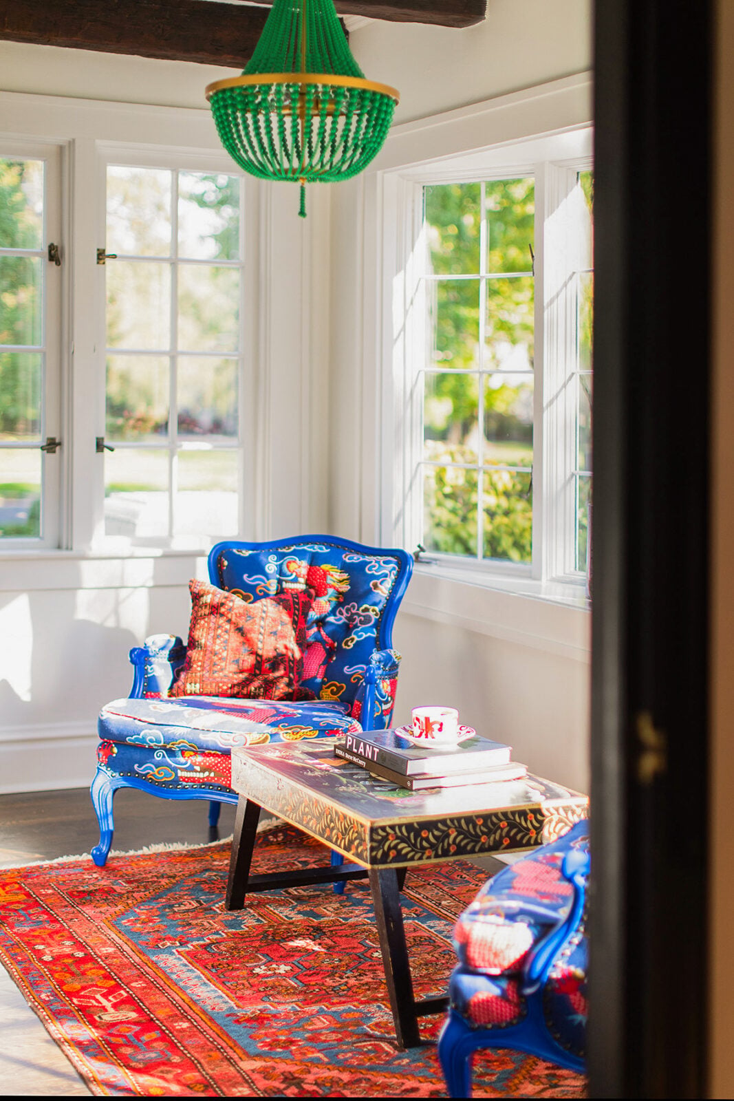 Bright sunroom with blue patterned armchairs, a colorful rug, a wooden coffee table with books, and a green chandelier. Large windows bring in natural light and garden views.