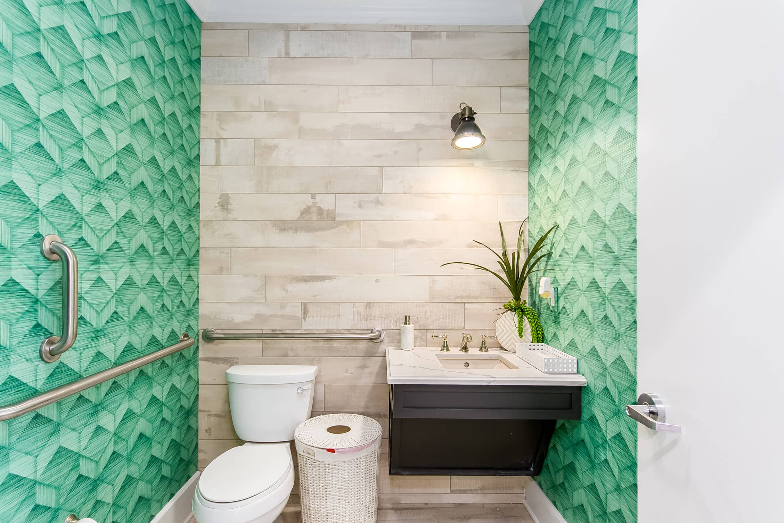 Small bathroom with green textured wallpaper, a white toilet, wicker laundry basket, wall-mounted sink with decor, and a single light fixture above.
