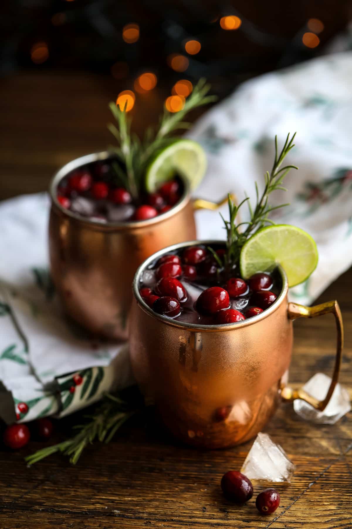Two copper mugs filled with a dark red drink, garnished with cranberries, lime slices, and rosemary, sit on a wooden table with ice cubes and a festive cloth—a cozy scene inspired by Crimson Design Group.