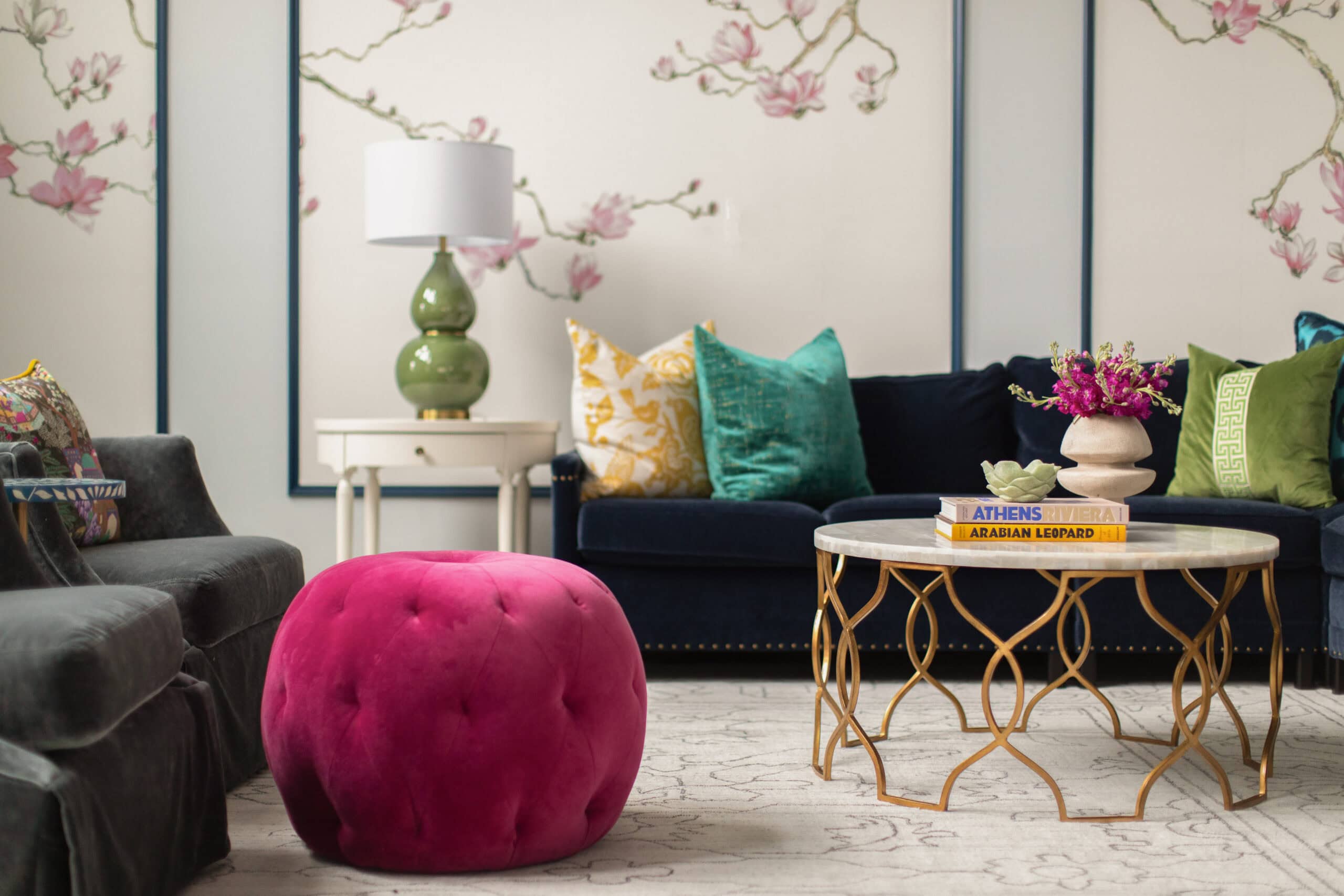 A living room with a dark sofa, colorful pillows, a round marble coffee table, a pink tufted ottoman, and floral wallpaper in the background.