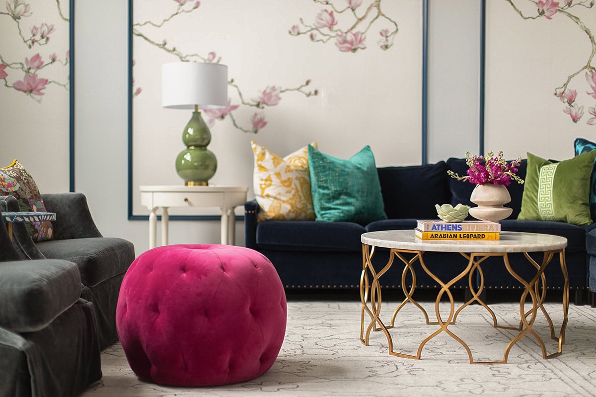 A living room with a navy sofa, assorted colorful pillows, a pink tufted ottoman, a round gold coffee table, and floral wallpaper in the background.