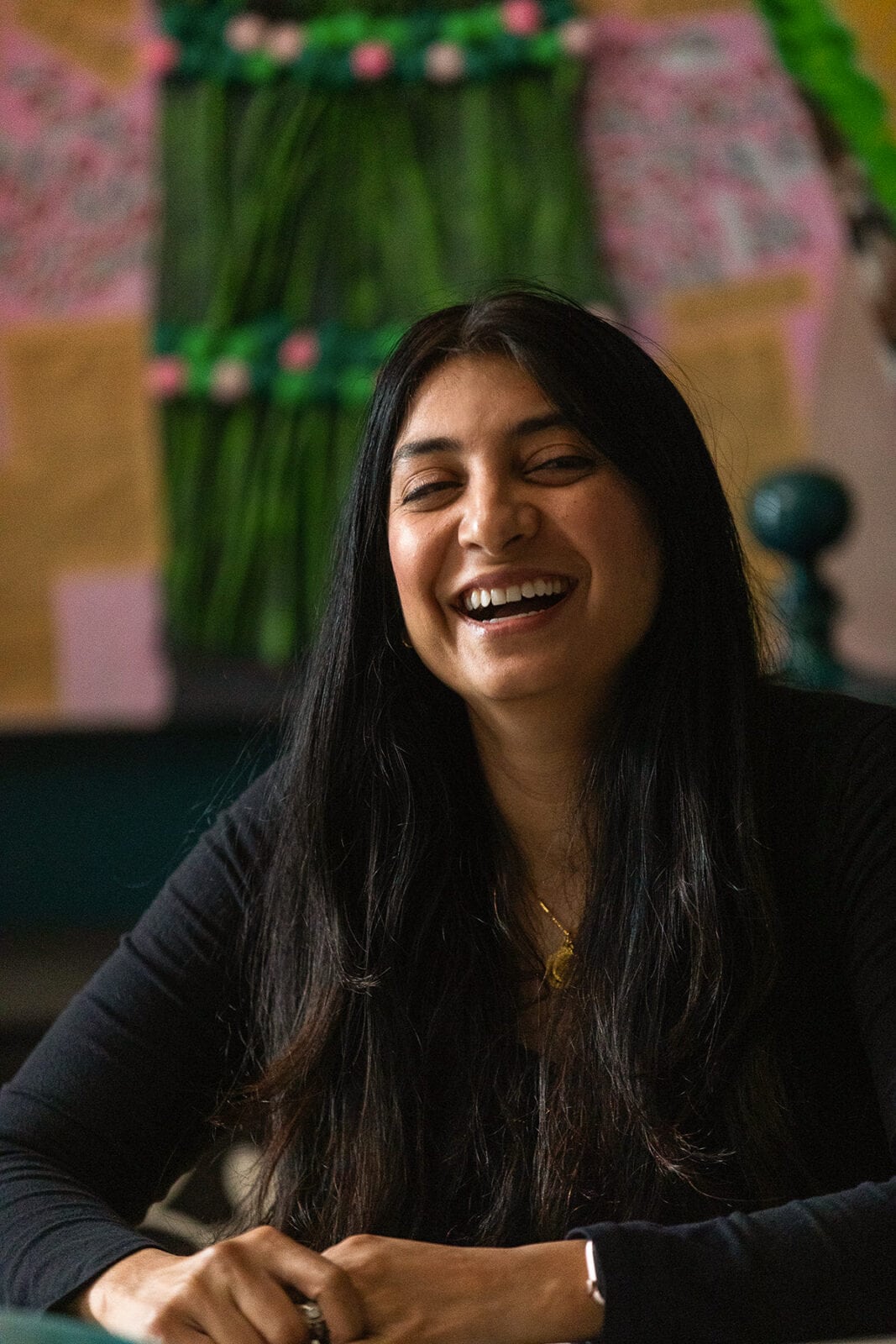 A candid, close-up portrait of Ashwini, a woman with long dark hair, laughing joyfully with a colorful, textured green and pink artwork blurred in the background.