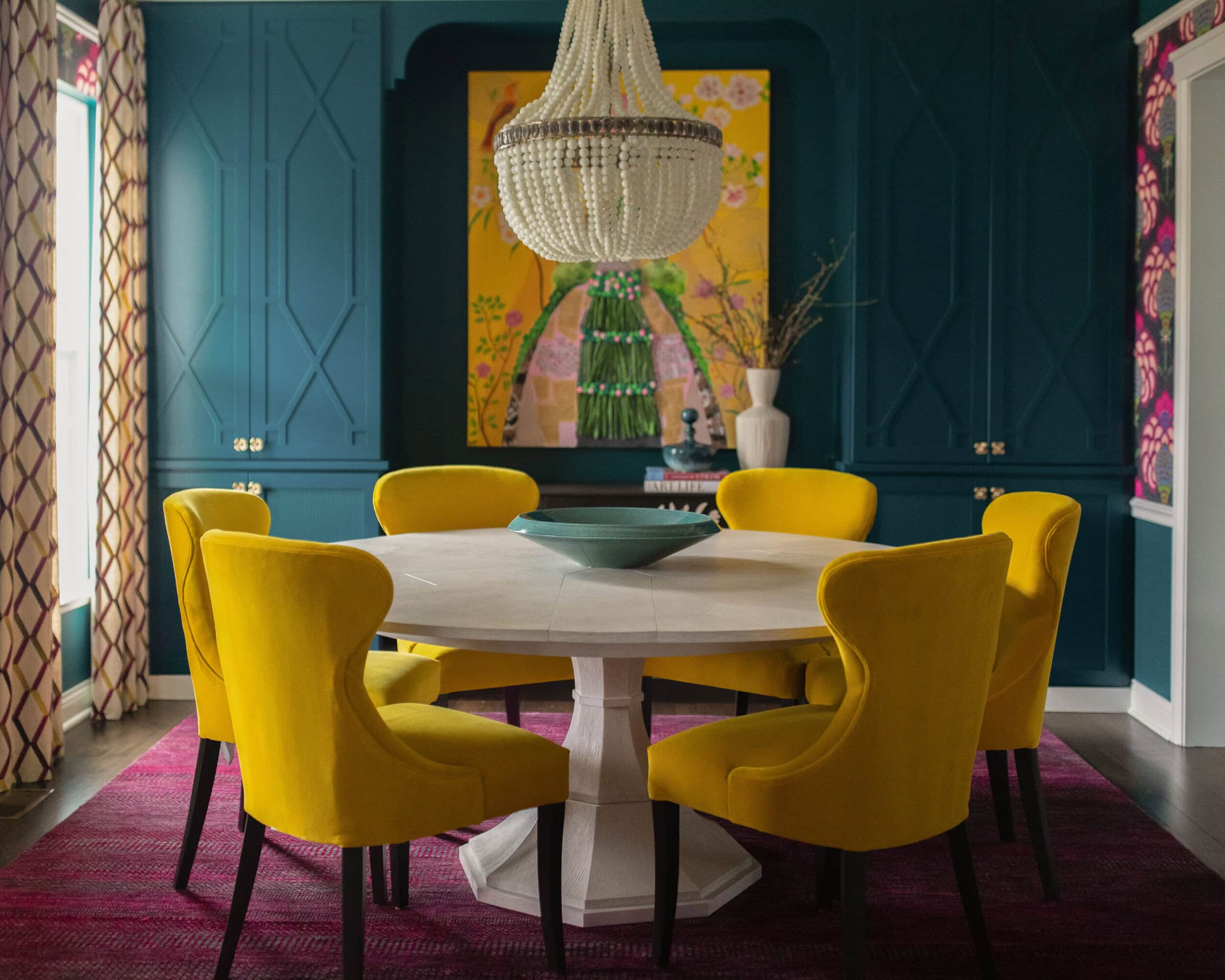 A vibrant dining room with dark teal cabinetry, six yellow wingback chairs around a round light-wood table, a large beaded chandelier, and colorful artwork on a yellow backdrop.