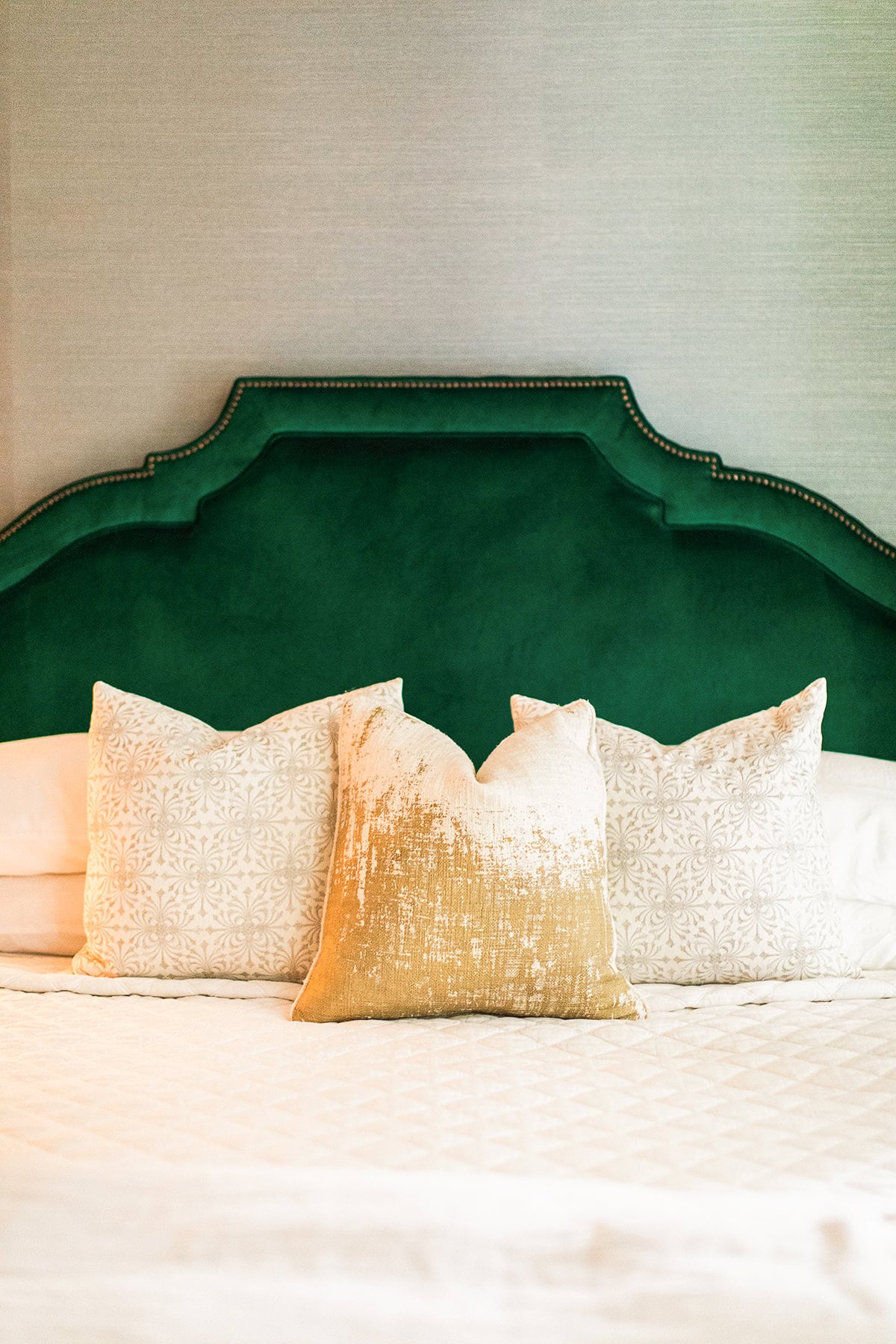 A neatly made bed with a green upholstered headboard, two white patterned pillows, and a gold decorative pillow in the center.