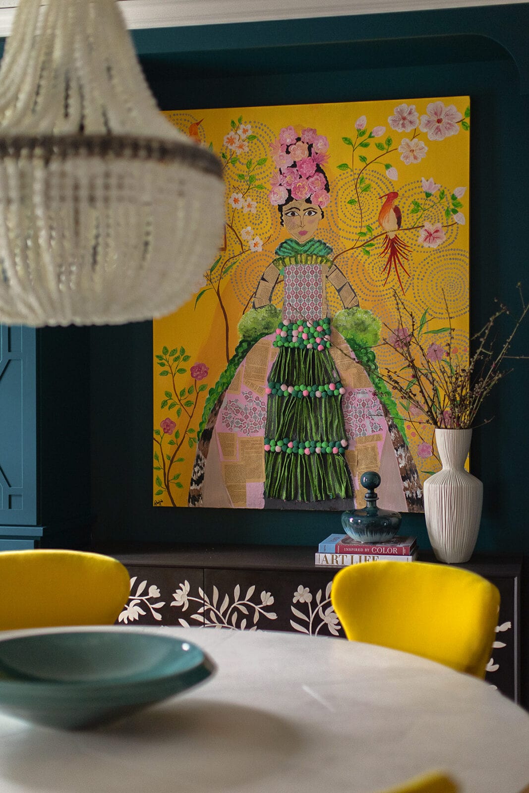A white beaded chandelier hangs over a dining table with yellow chairs; behind is a colorful portrait of a woman with flowers on a yellow background and a sideboard with decor items.