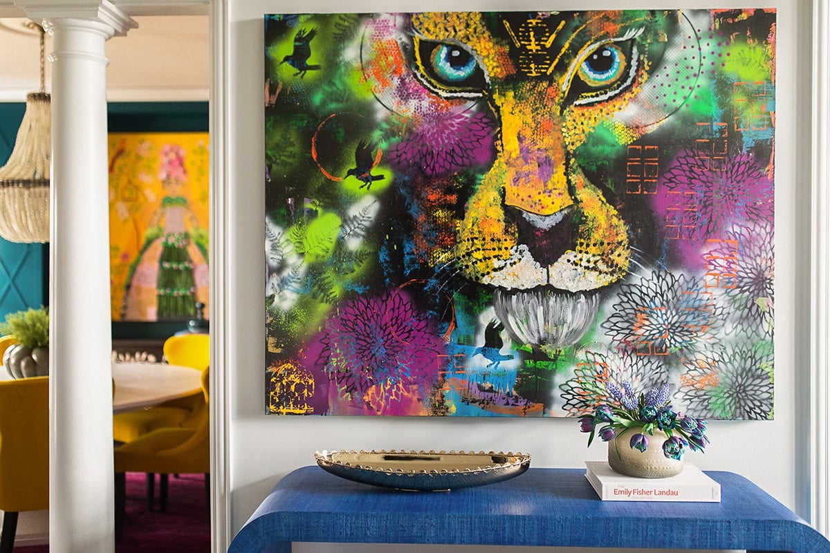 A vibrant, multi-colored graffiti-style painting of a lion’s face hangs above a blue textured console table with a gold bowl and purple tulips. In the background, a dining room with yellow chairs is visible through a doorway.