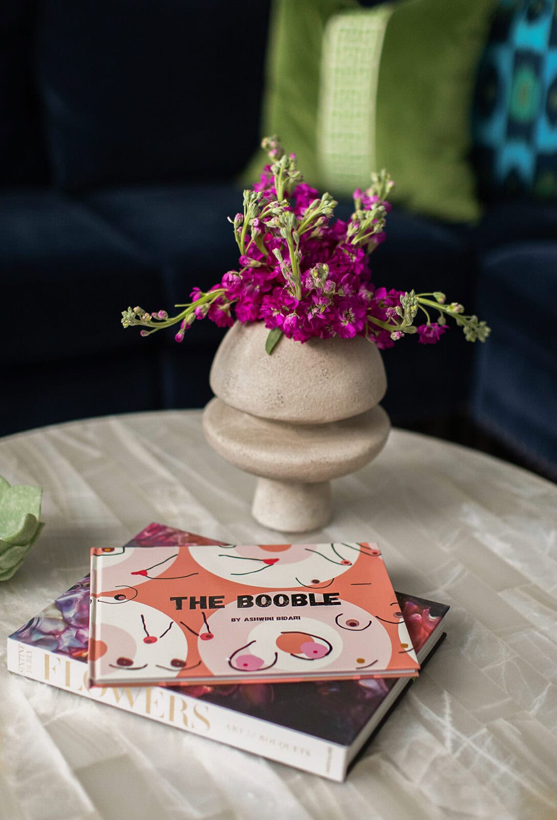 A round beige vase with pink flowers sits on a table next to two books, one titled "The Booble" with an illustrated cover and another titled "Flowers.