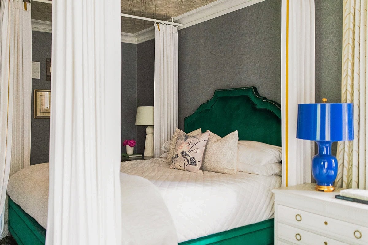 A bedroom features a green velvet canopy bed with white curtains, neutral bedding, decorative pillows, a white nightstand, and a blue lamp.