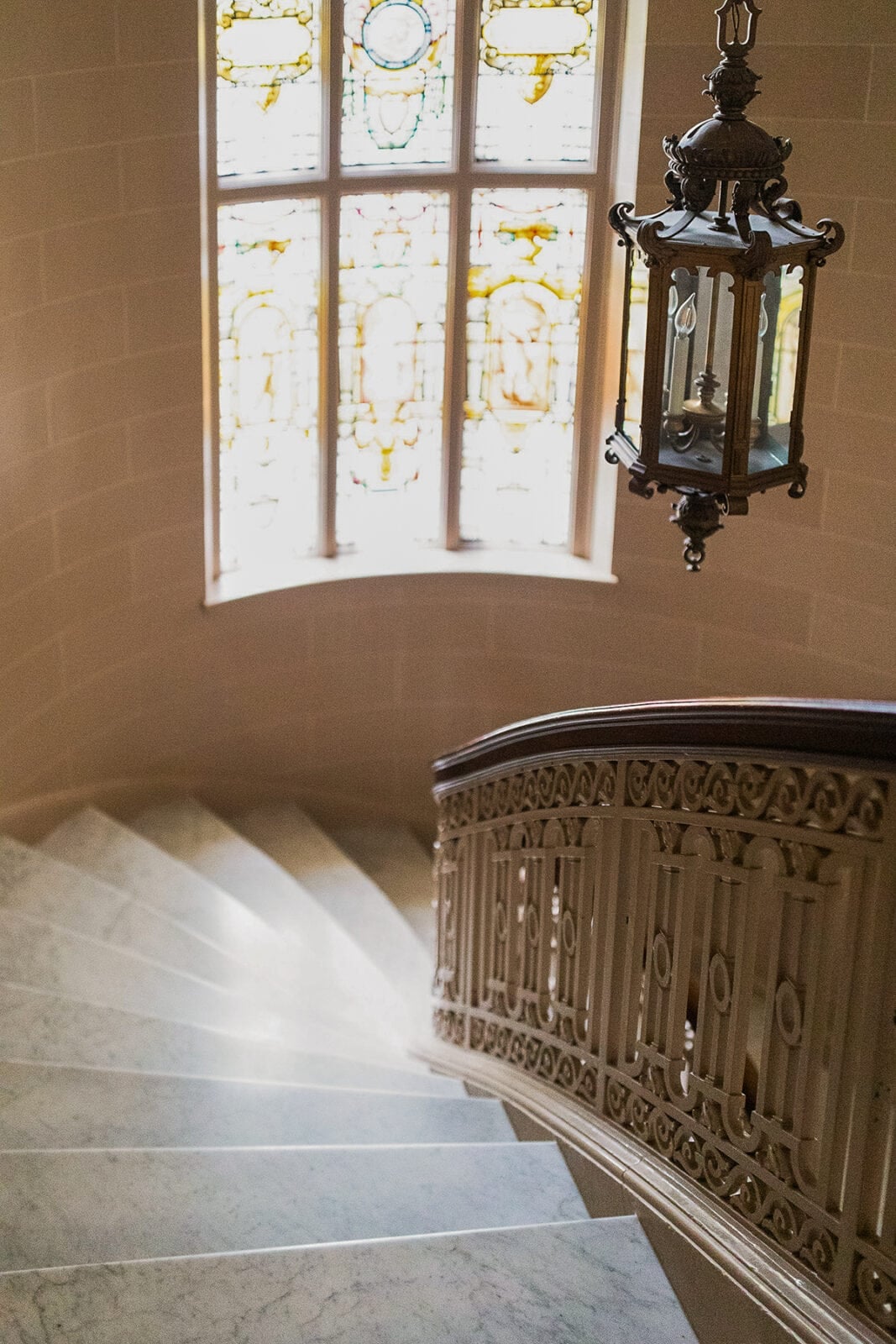 Curved marble staircase with ornate railing, leading down toward a large window with stained glass and a decorative hanging lantern.