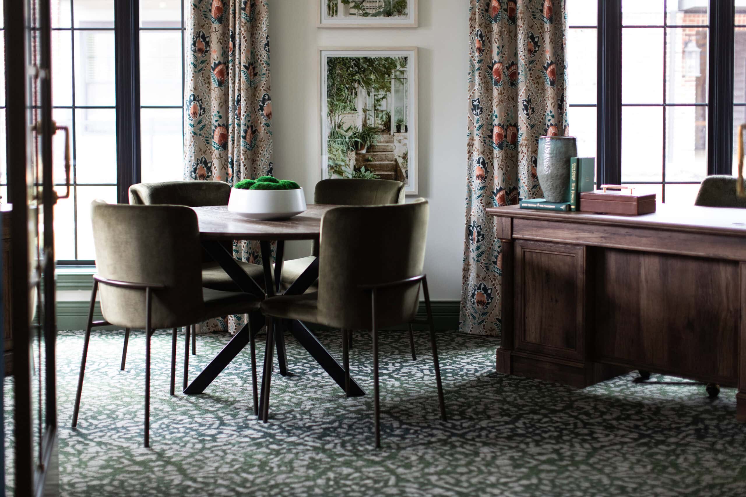 Sophisticated commercial office interior featuring a round walnut meeting table, olive green velvet chairs, patterned moss-green carpeting, and a custom wooden executive desk with botanical artwork.