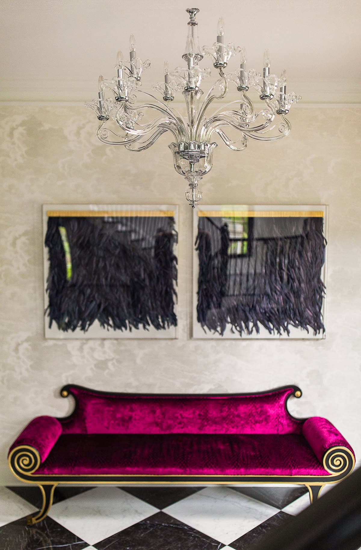 A glass chandelier hangs above a magenta velvet sofa with gold accents, set against a wall with two abstract black artworks and a black-and-white checkered floor.