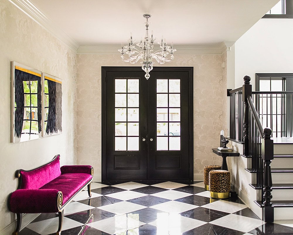 A foyer with black double doors, a glass chandelier, black and white checkered floor, magenta velvet bench, two paintings, and a staircase with black railing.