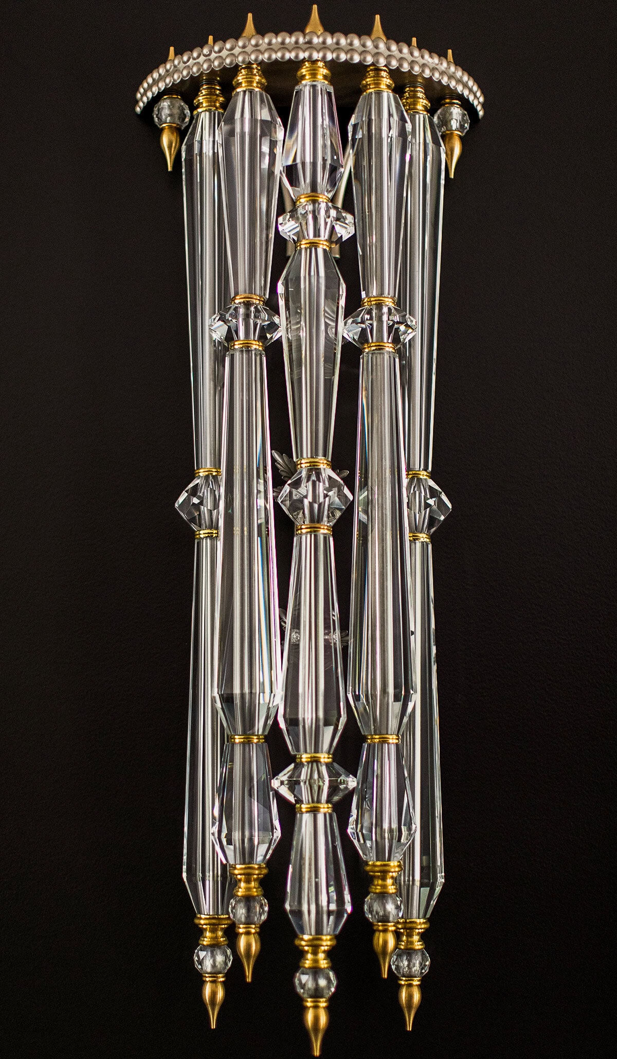 A decorative wall sconce featuring vertically aligned crystal rods with gold accents, set against a black background.