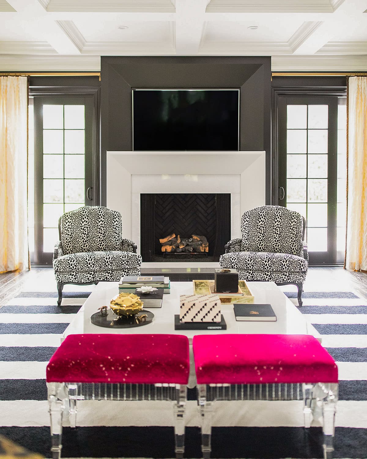 A modern living room with two patterned armchairs, a fireplace, a wall-mounted TV, a striped rug, a coffee table, and two clear benches with bright pink cushions.