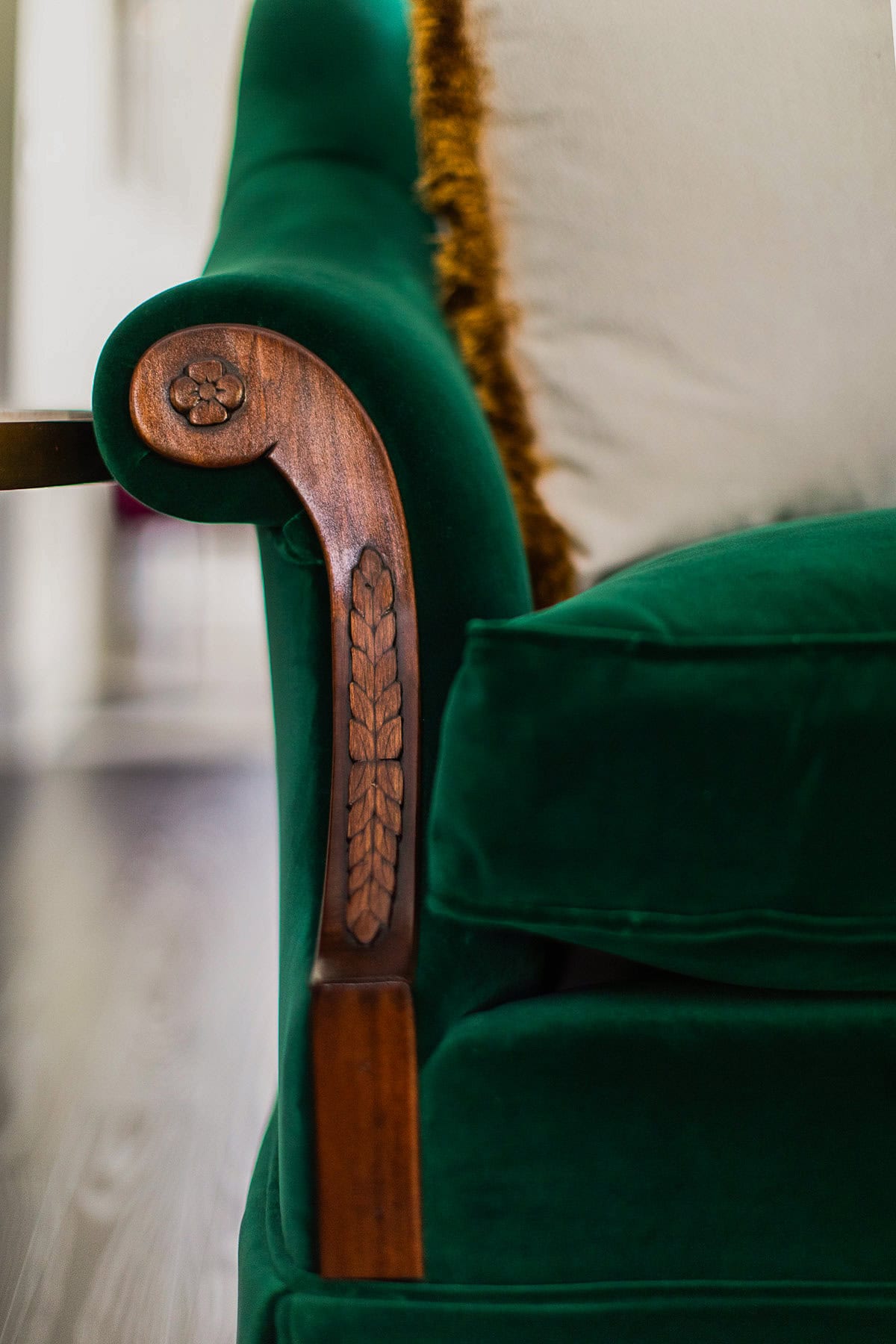 Close-up of a green velvet armchair with carved wooden armrest details and a white pillow with a textured edge.