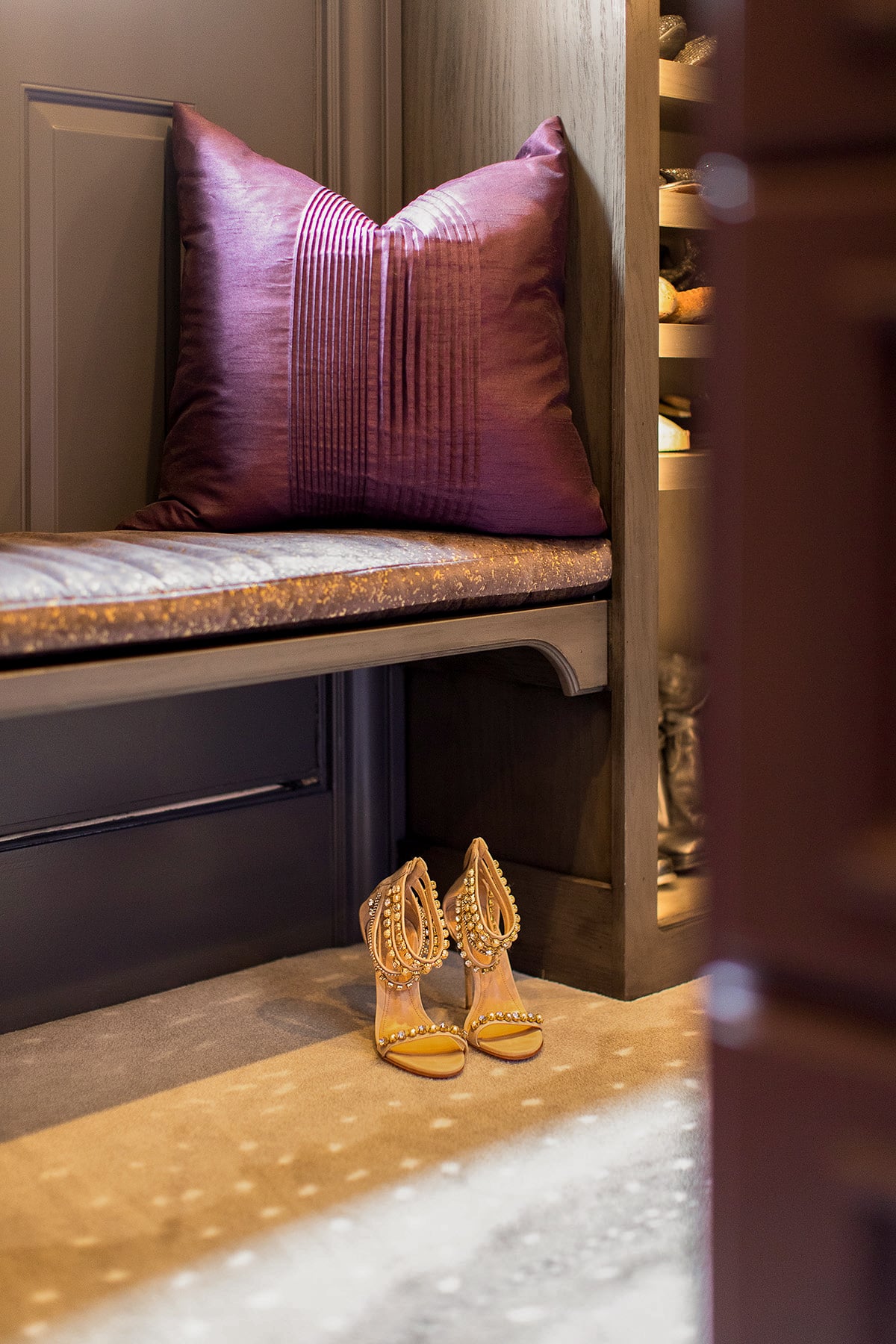 A pair of embellished high-heeled sandals sits on the floor in front of a cushioned bench with a purple pillow.