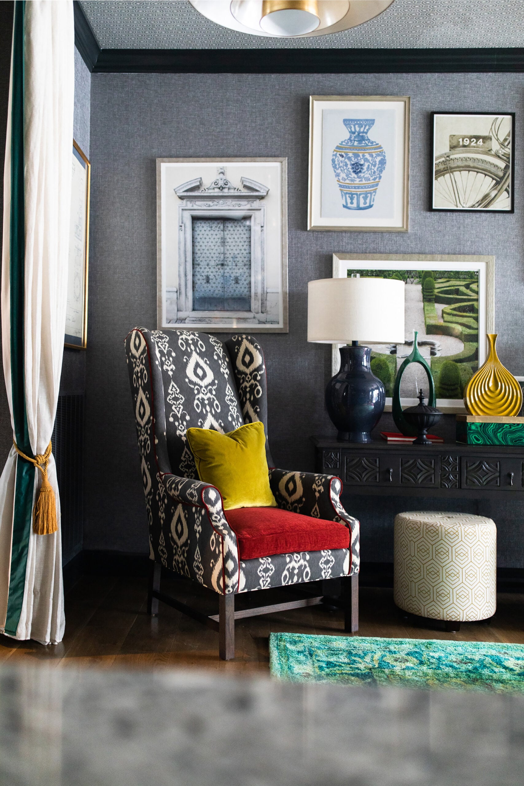 A patterned armchair with a yellow pillow sits beside a modern table, lamp, and decor; framed art hangs on a gray wall above a green rug and pouf.