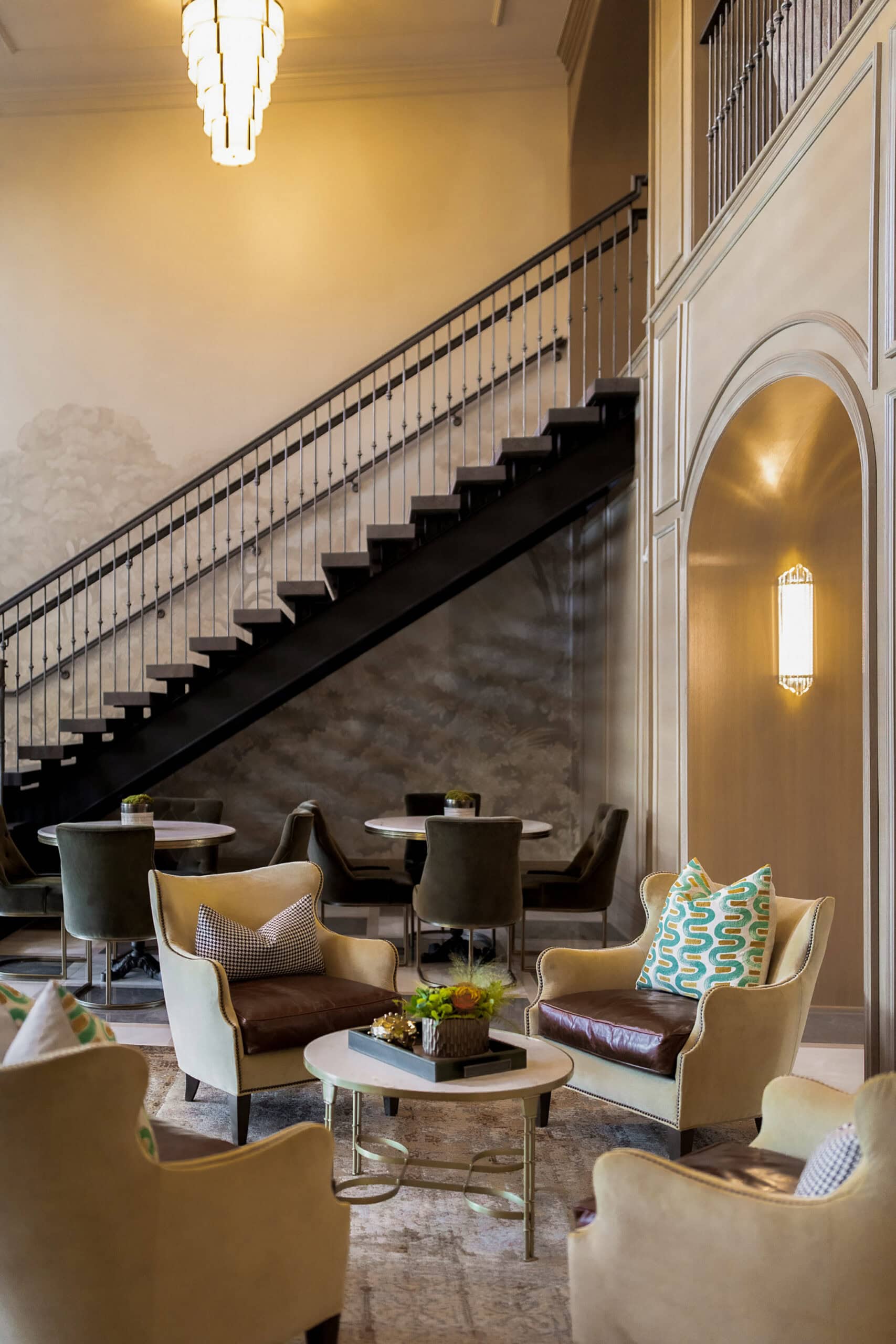 A modern lounge area with beige armchairs, patterned cushions, small round tables, and a black metal staircase against a light-colored wall.