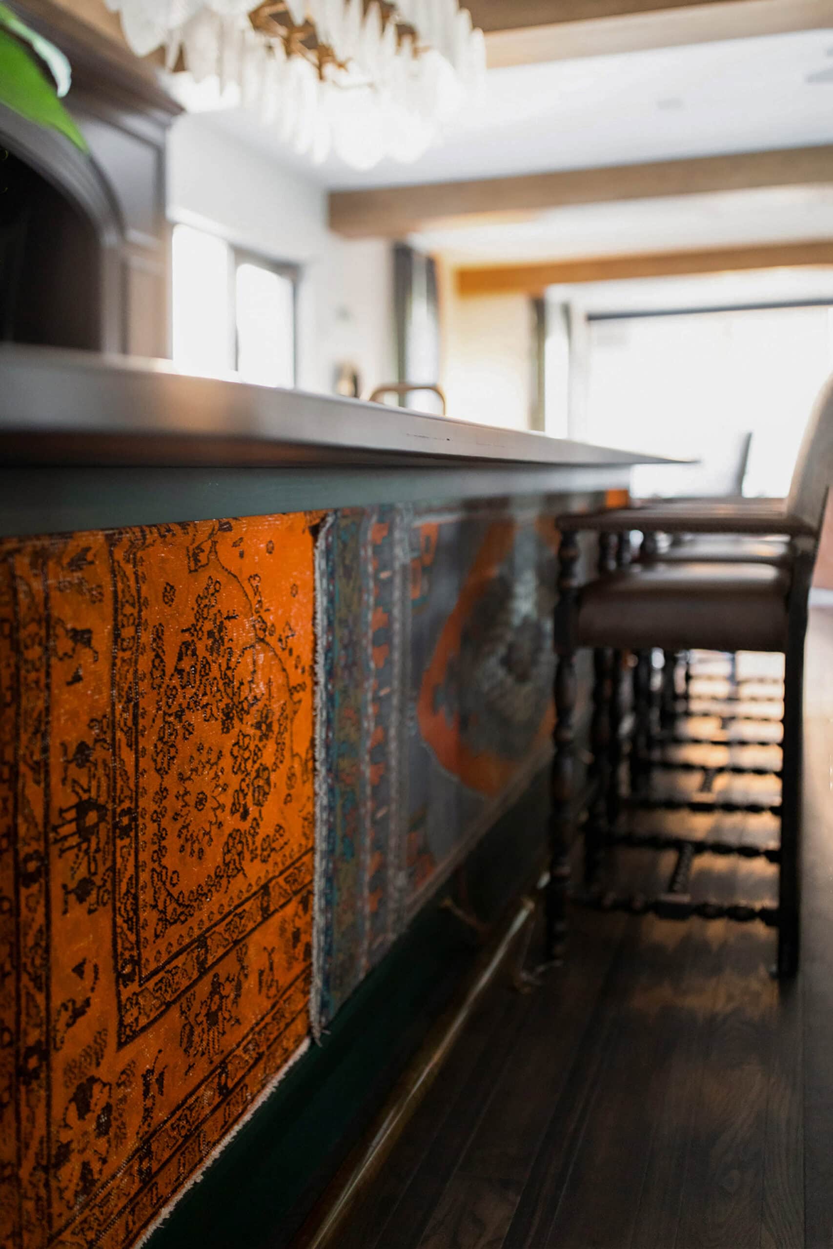 A close-up of a bar with upholstered orange patterned panels, wooden chairs, and sunlight streaming in from large windows in the background.