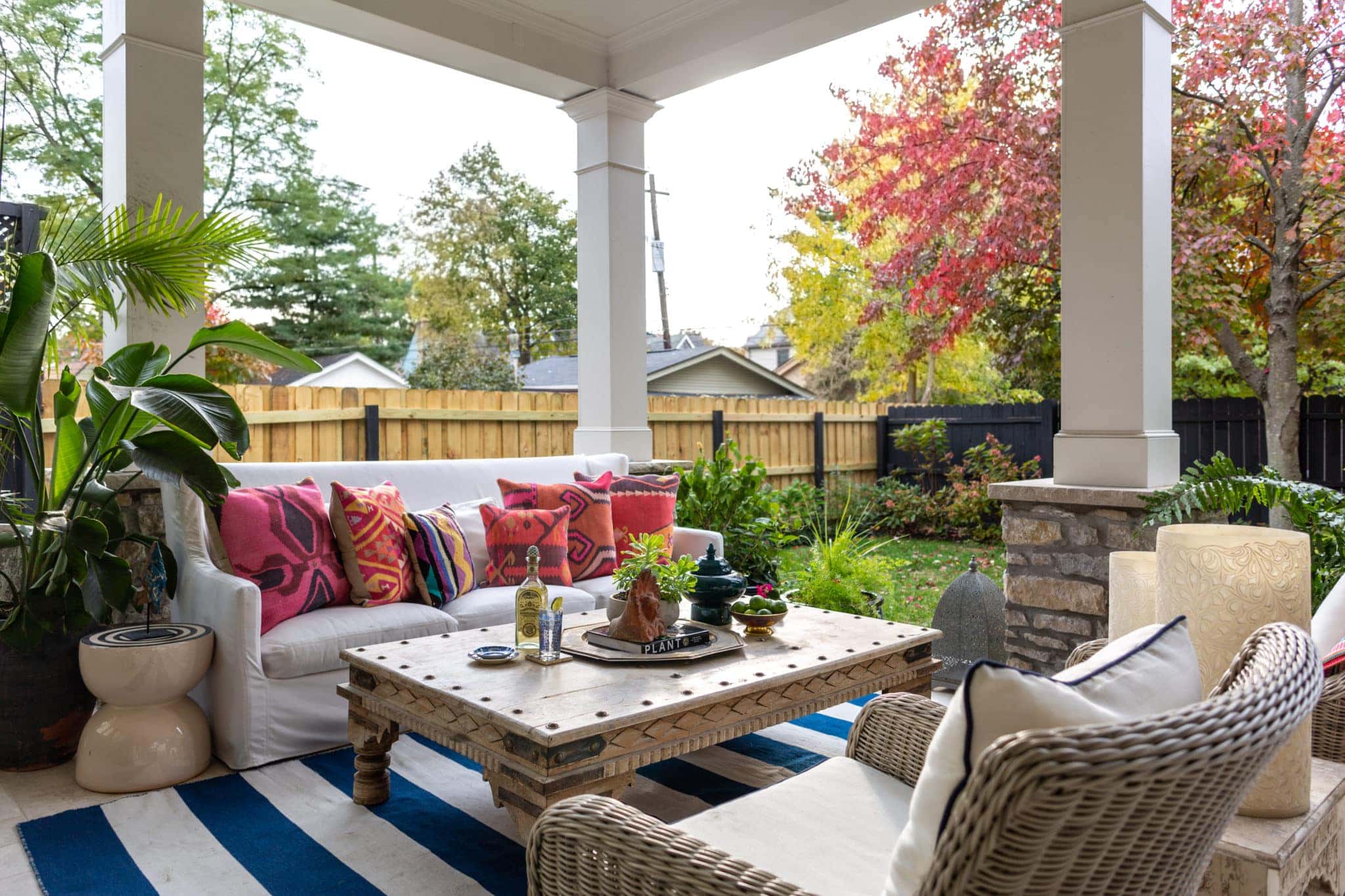 A covered patio designed by Crimson Design Group features a white sofa with colorful cushions, wicker chairs, and a wooden coffee table, all set beside a fenced backyard adorned with autumn-colored trees.