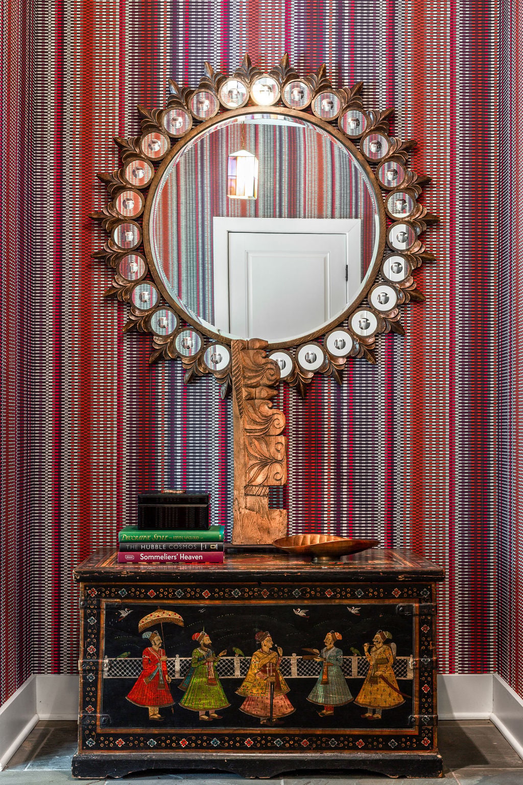 Decorative dresser with painted figures, books, and gold decor sits below a round mirror with ornate frame, set against red and purple striped wallpaper. A white door is reflected in the mirror.