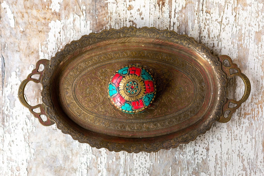 An ornate, oval brass tray with handles holds a round, colorful, mosaic-decorated box on a distressed wooden surface.