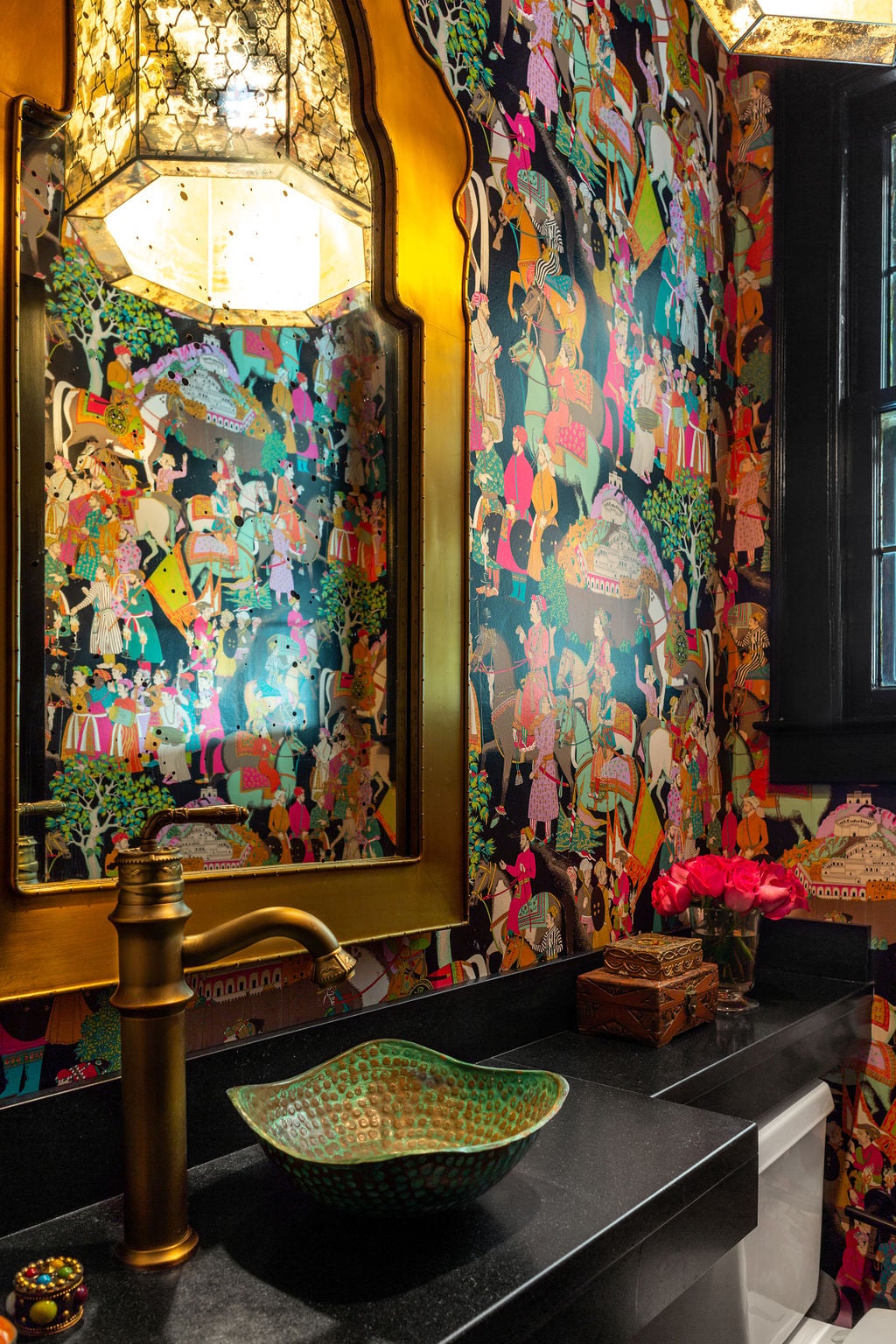 Bathroom with colorful, intricate wallpaper, a gold-framed mirror, brass faucet, green vessel sink, black countertop, and decorative items, including pink flowers and a small patterned box.