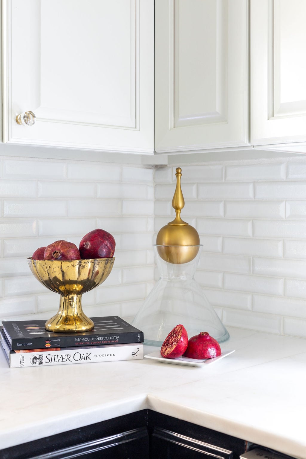 A kitchen countertop with pomegranates in a gold bowl, a glass decanter with a gold top, three cookbooks, and a white plate holding a sliced pomegranate.