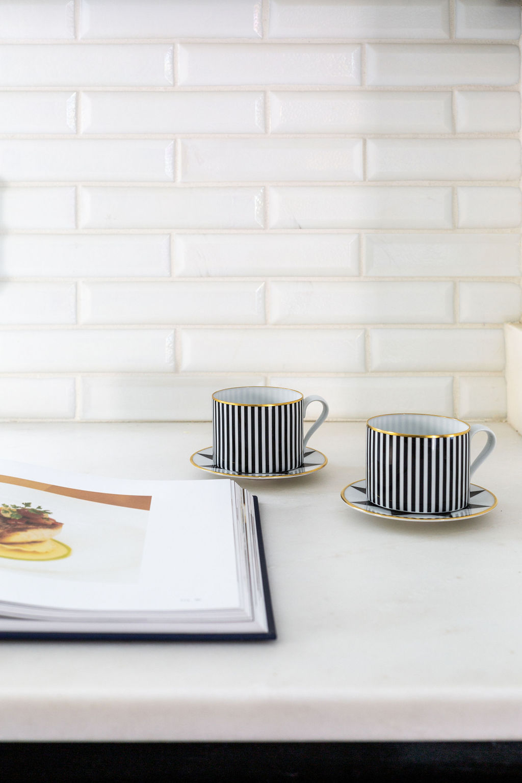 Two black and white striped teacups with saucers sit on a white countertop next to an open cookbook, against a white tiled backsplash.