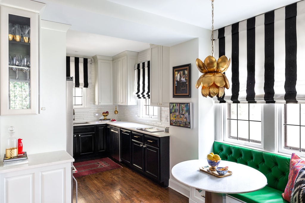 A modern kitchen with black and white striped window shades, black lower cabinets, white upper cabinets, and a green tufted bench next to a round table.