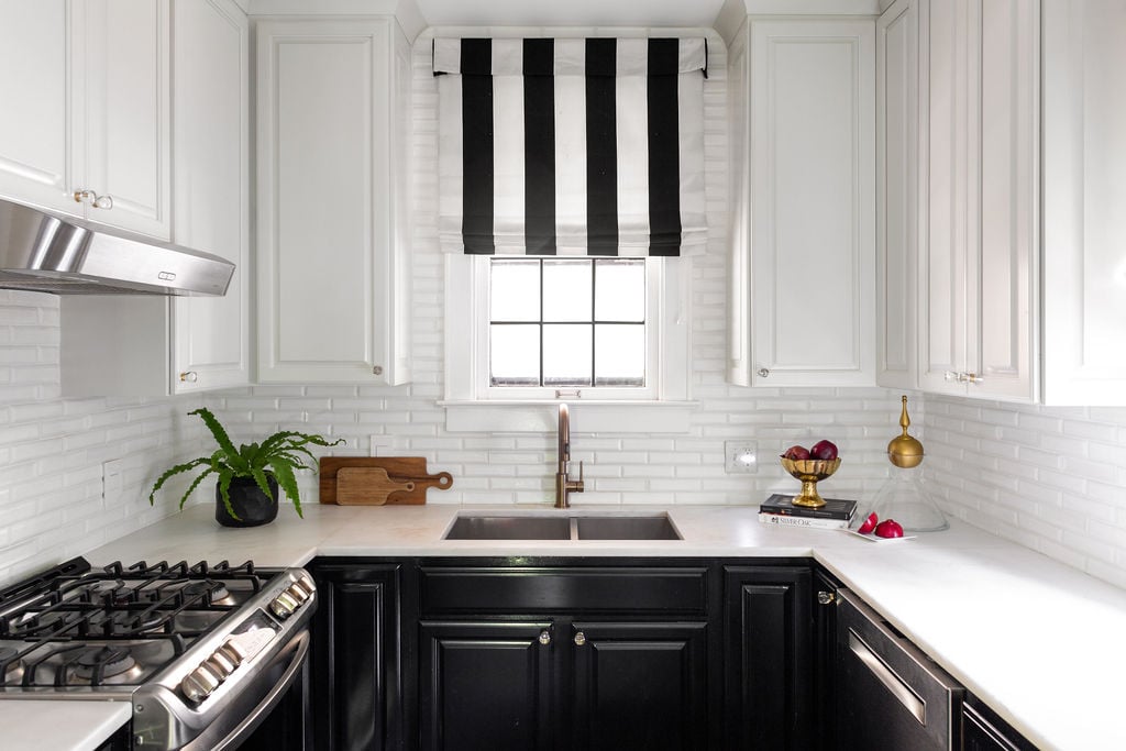 A modern kitchen with black lower cabinets, white upper cabinets, a stainless steel stove, white subway tile backsplash, and a window with a black-and-white striped shade.