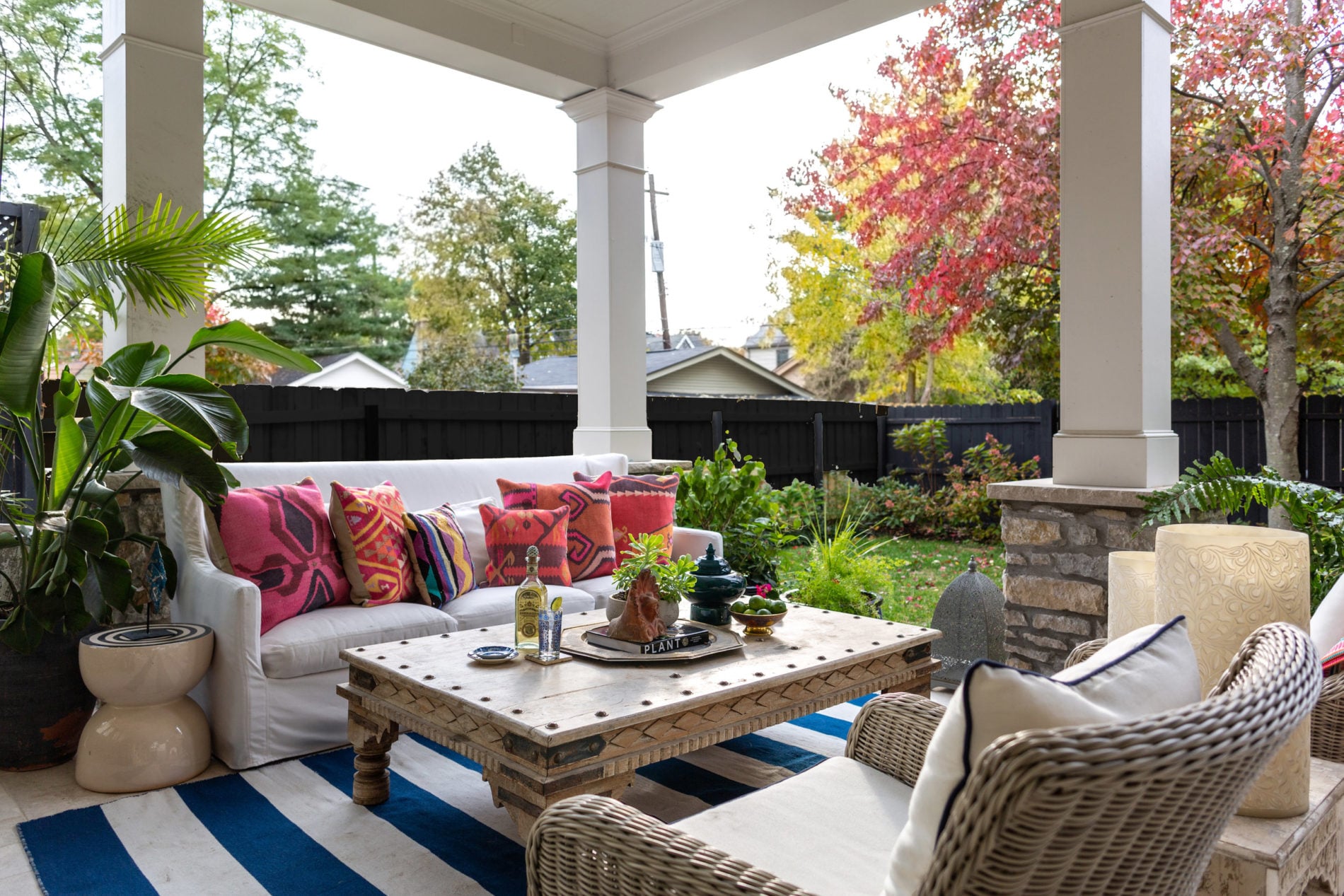 A covered patio designed by Crimson Design Group features a white sofa, colorful pillows, a wooden coffee table, wicker chairs, a striped rug, and a stunning garden view with autumn trees.
