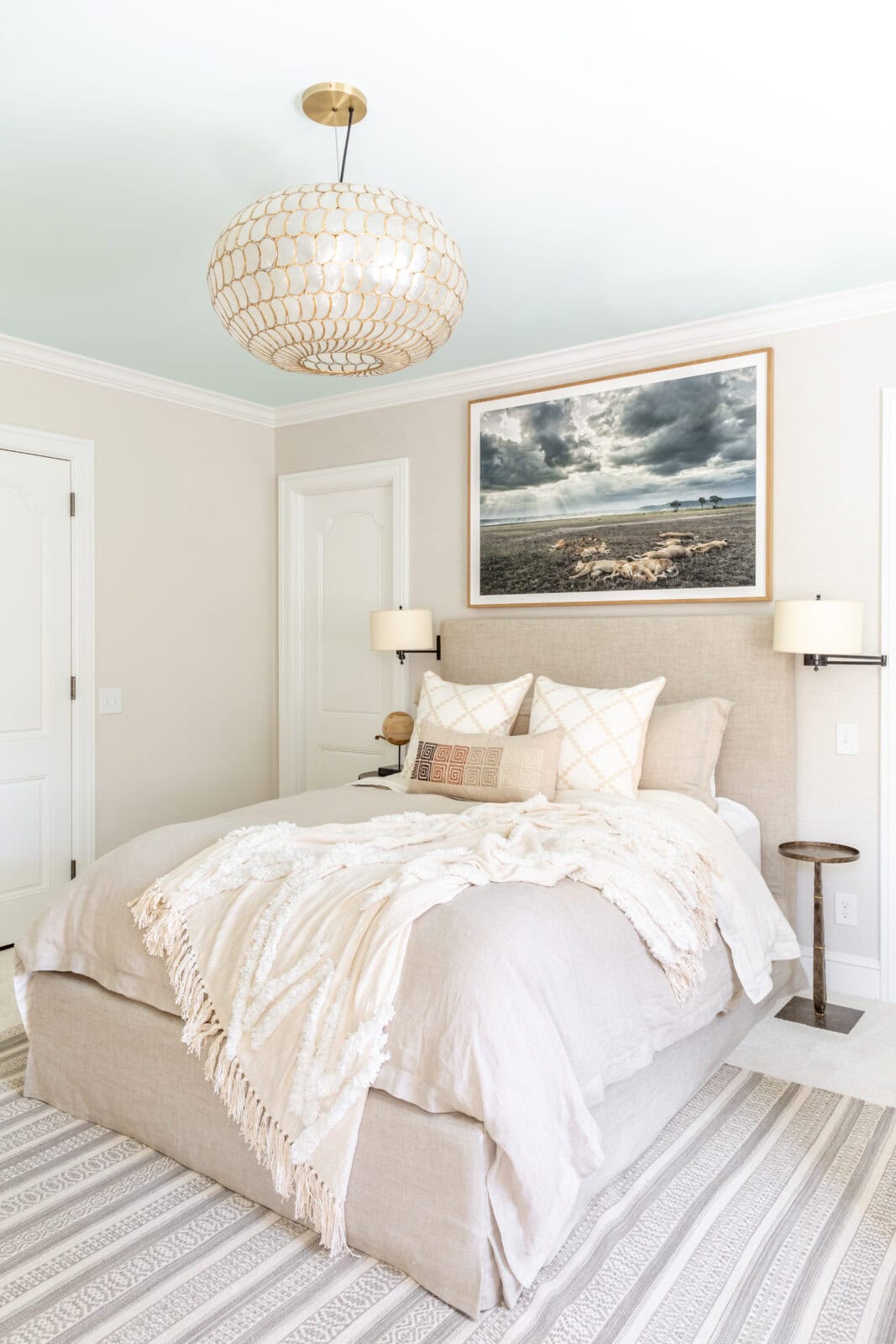Neutral-toned bedroom with a beige upholstered bed, layered blankets, striped rug, two wall-mounted lamps, and a large framed ocean photograph above the headboard.
