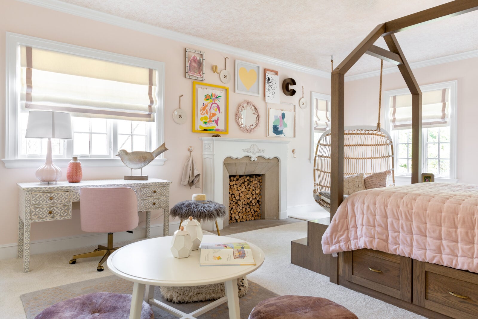 A pastel pink bedroom with a canopy bed, desk, hanging chair, fireplace with stacked logs, and children’s decor, including wall art and toys.
