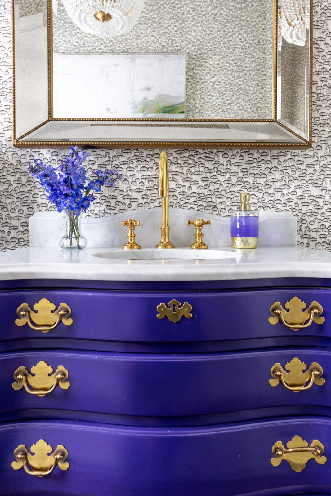 A bathroom vanity with a purple and gold dresser, gold faucet, blue flowers in a vase, soap dispenser, and a large mirror above.