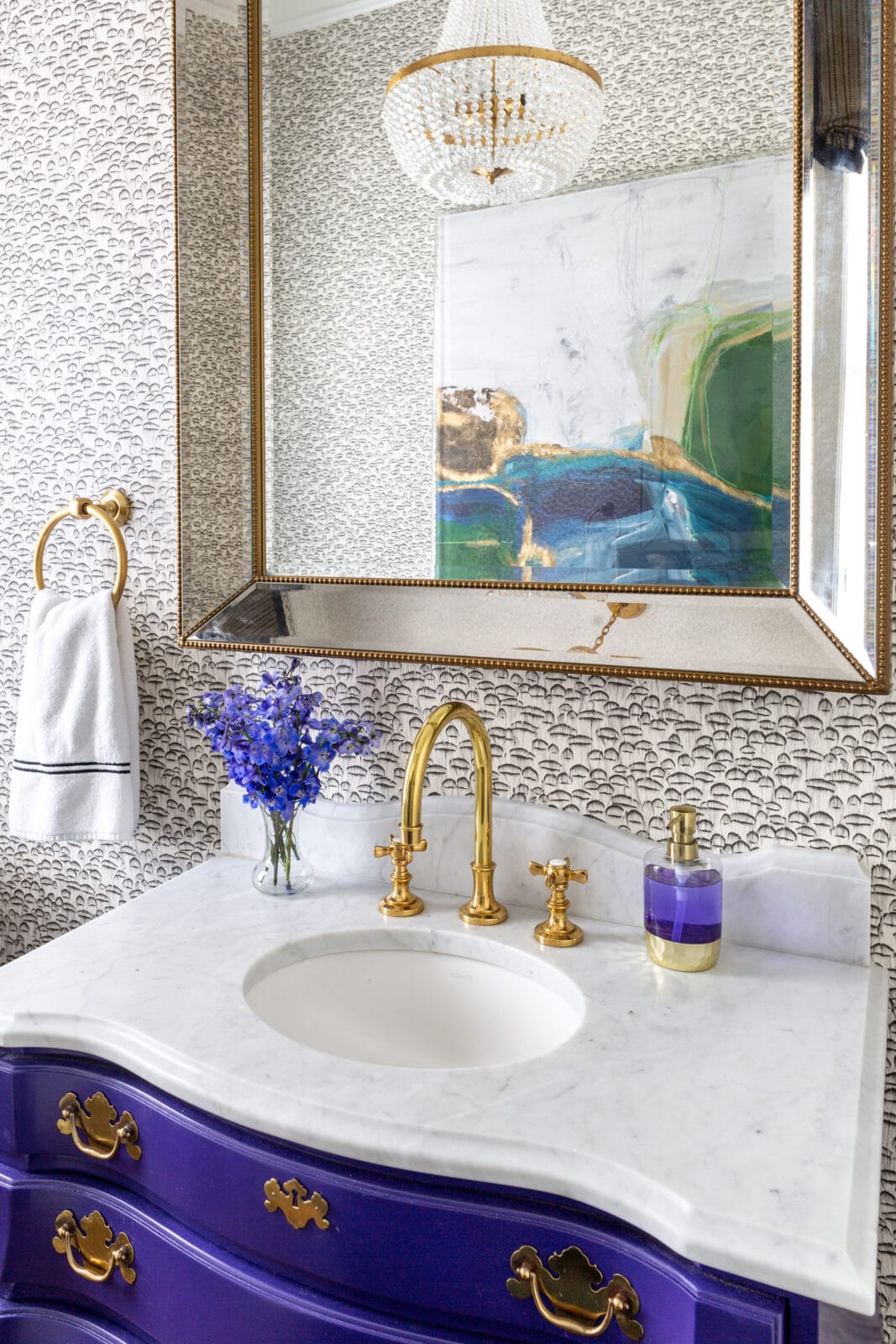 A bathroom vanity with a white marble countertop, gold faucet, purple drawers, a framed mirror, and a vase of purple flowers. Abstract art and a chandelier are visible.