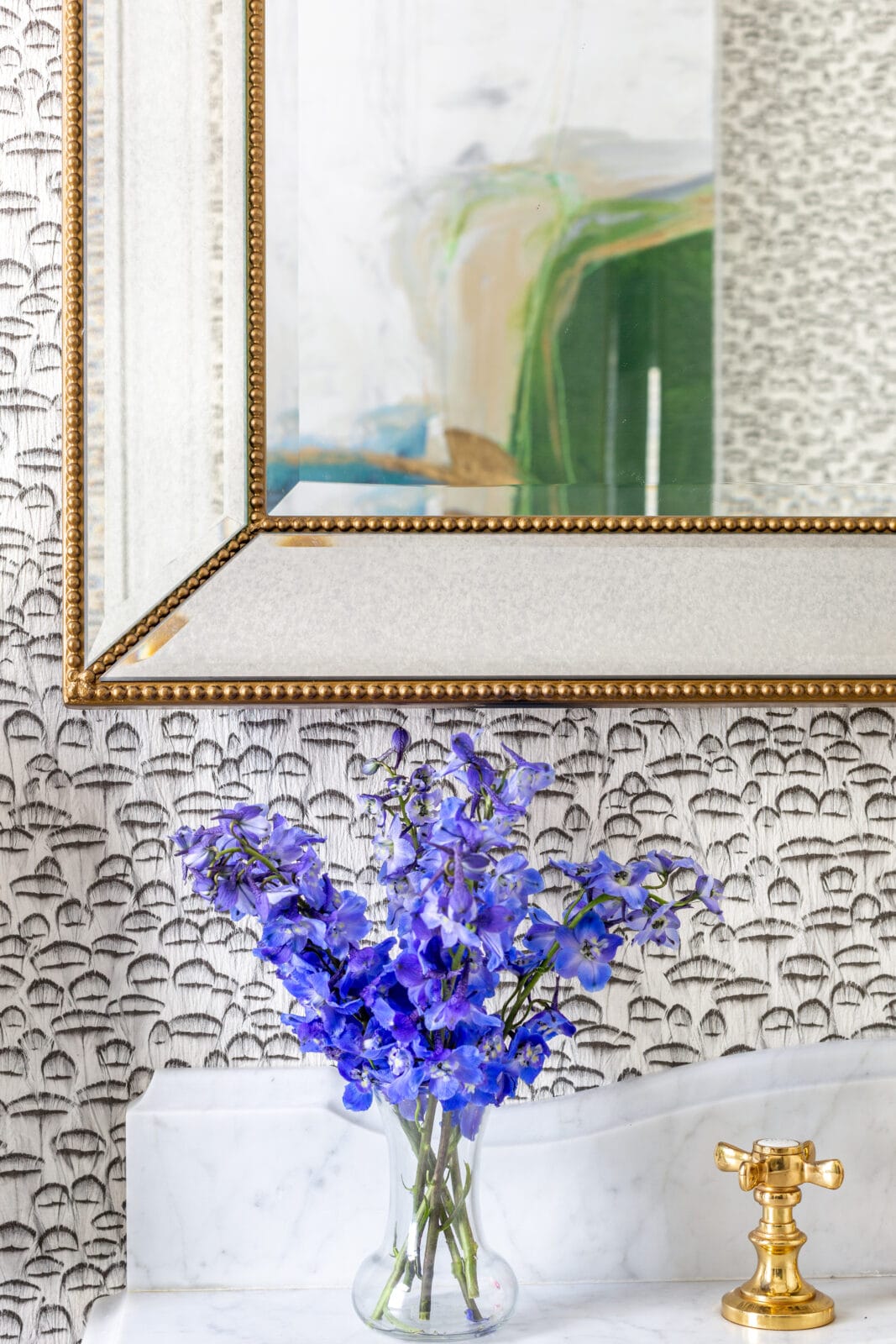 A vase with purple flowers sits on a marble countertop beneath a large mirror with a gold frame and a gold faucet, against patterned wallpaper.