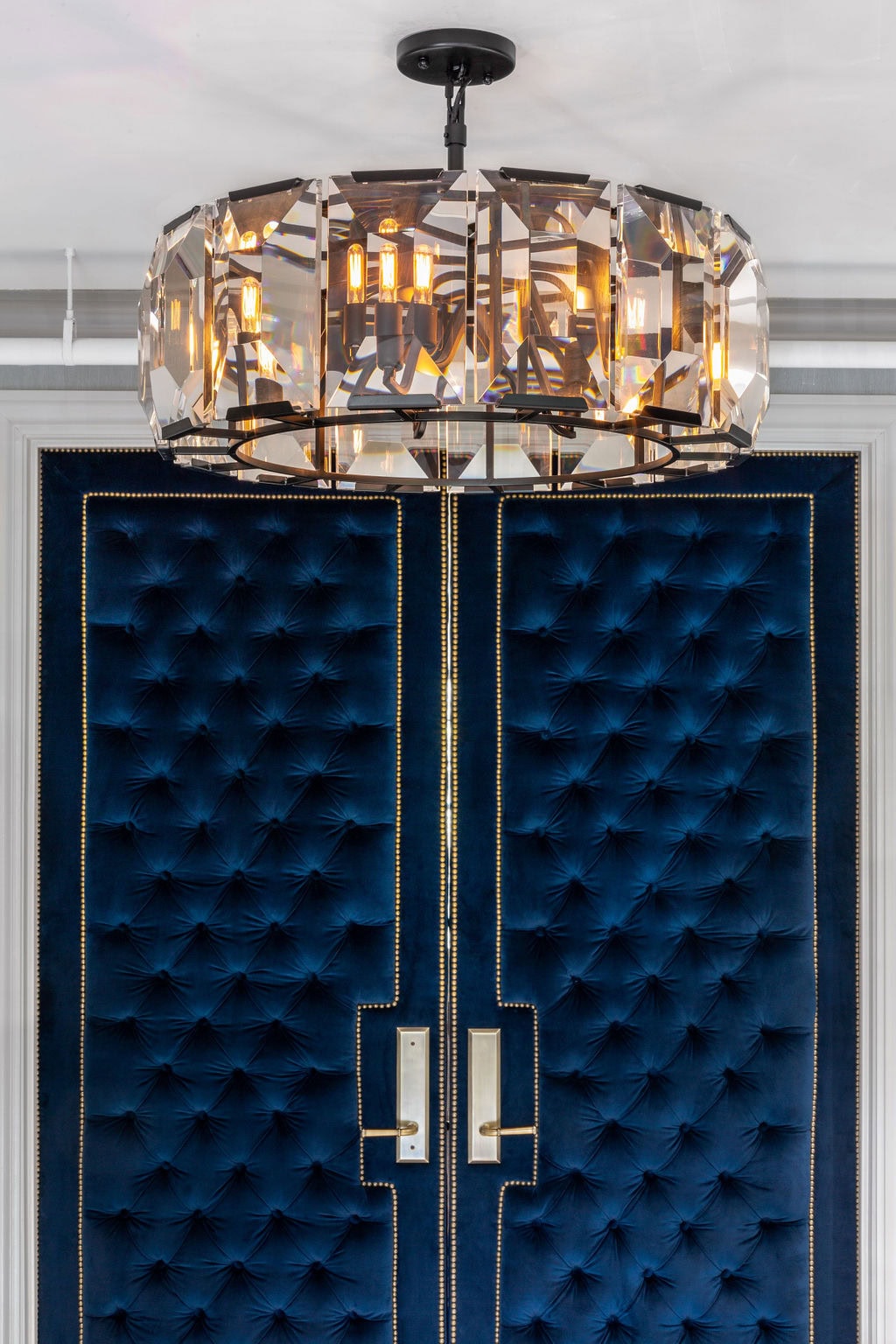 A geometric crystal chandelier hangs above double doors upholstered in deep blue tufted fabric with gold trim and handles.