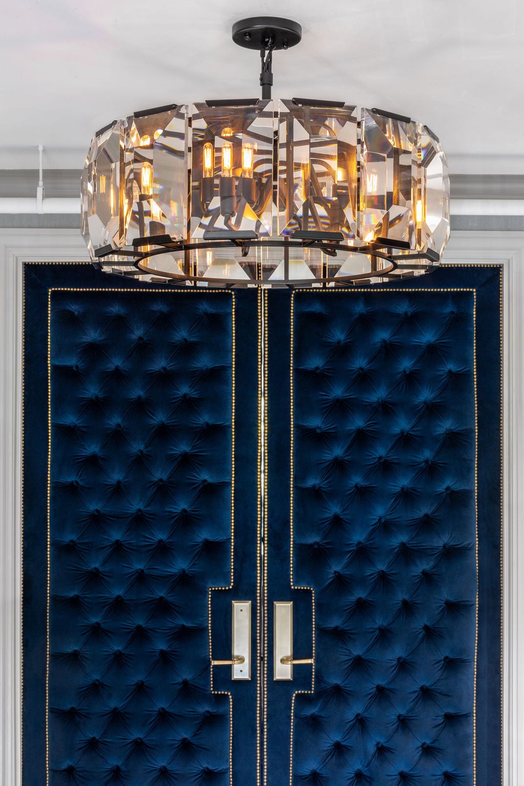 A crystal chandelier with exposed bulbs hangs above double doors upholstered in deep blue tufted fabric with gold trim and metallic handles.