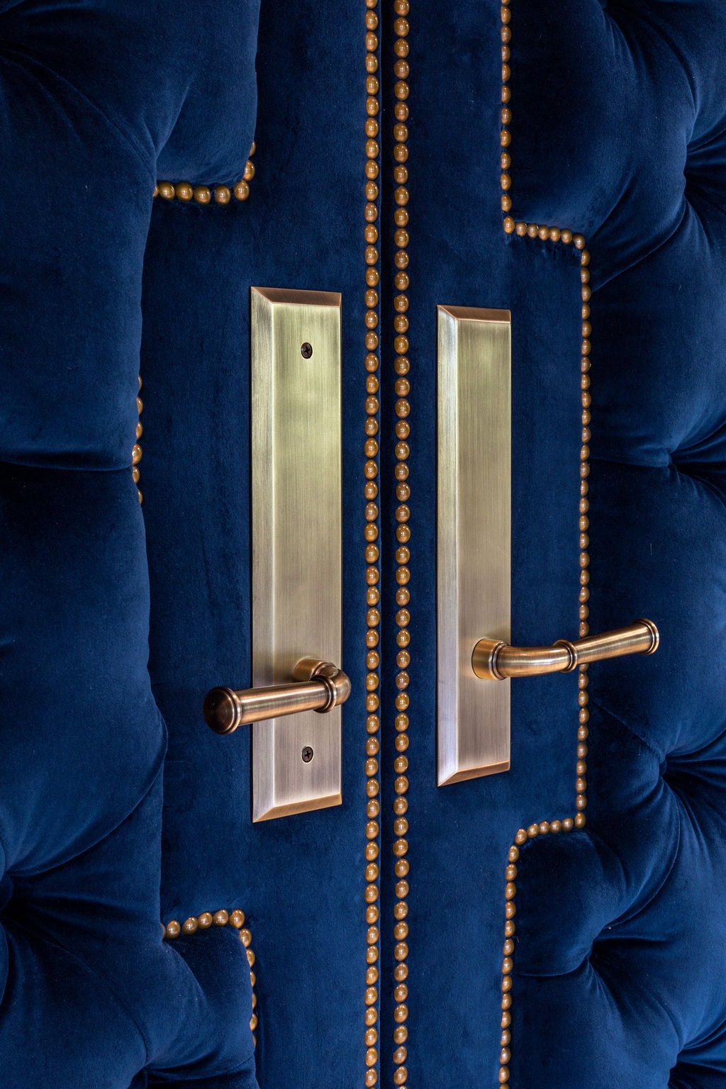 Close-up of two gold door handles on a tufted, deep blue velvet door with gold decorative studs outlining the upholstery.
