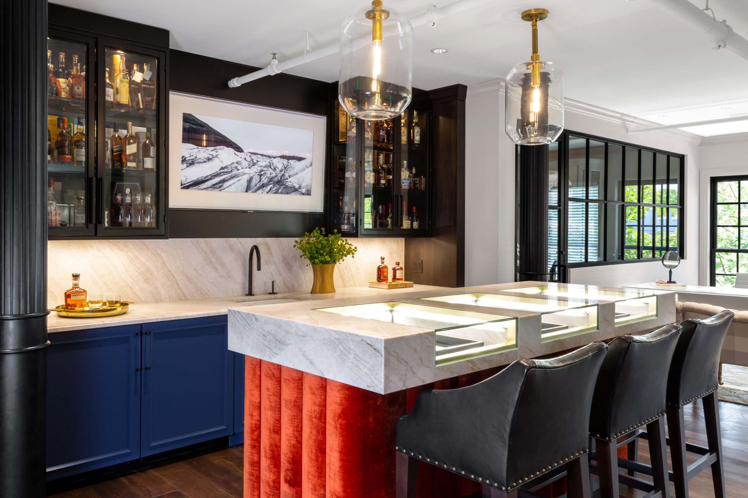 Modern home bar with marble counter, red velvet bar front, black leather stools, glass pendant lights, and shelves stocked with bottles; framed landscape photo on wall above sink.