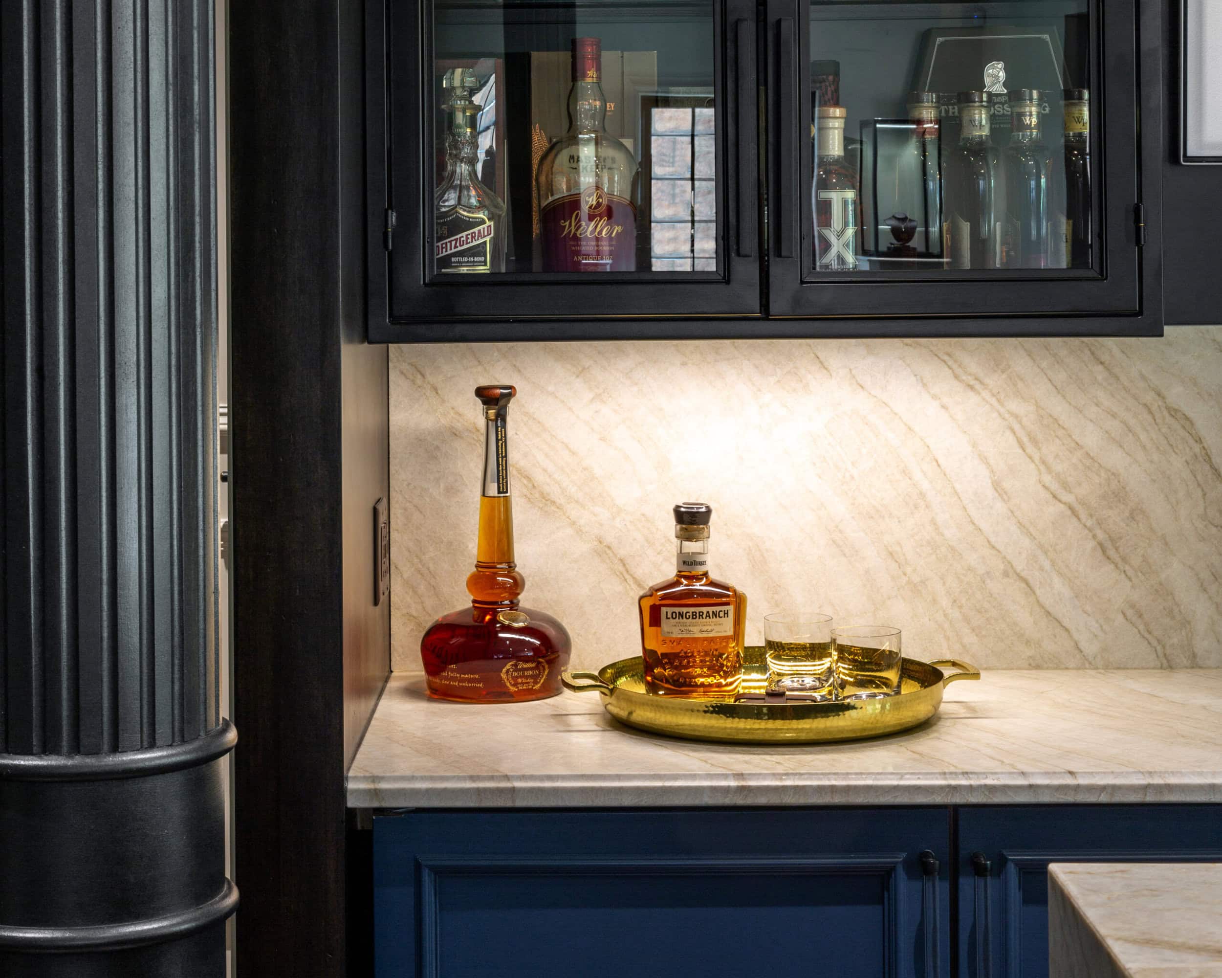 A home bar setup with bottles of liquor, two glasses, and a decanter arranged on a gold tray on a marble countertop beneath glass-front cabinets.